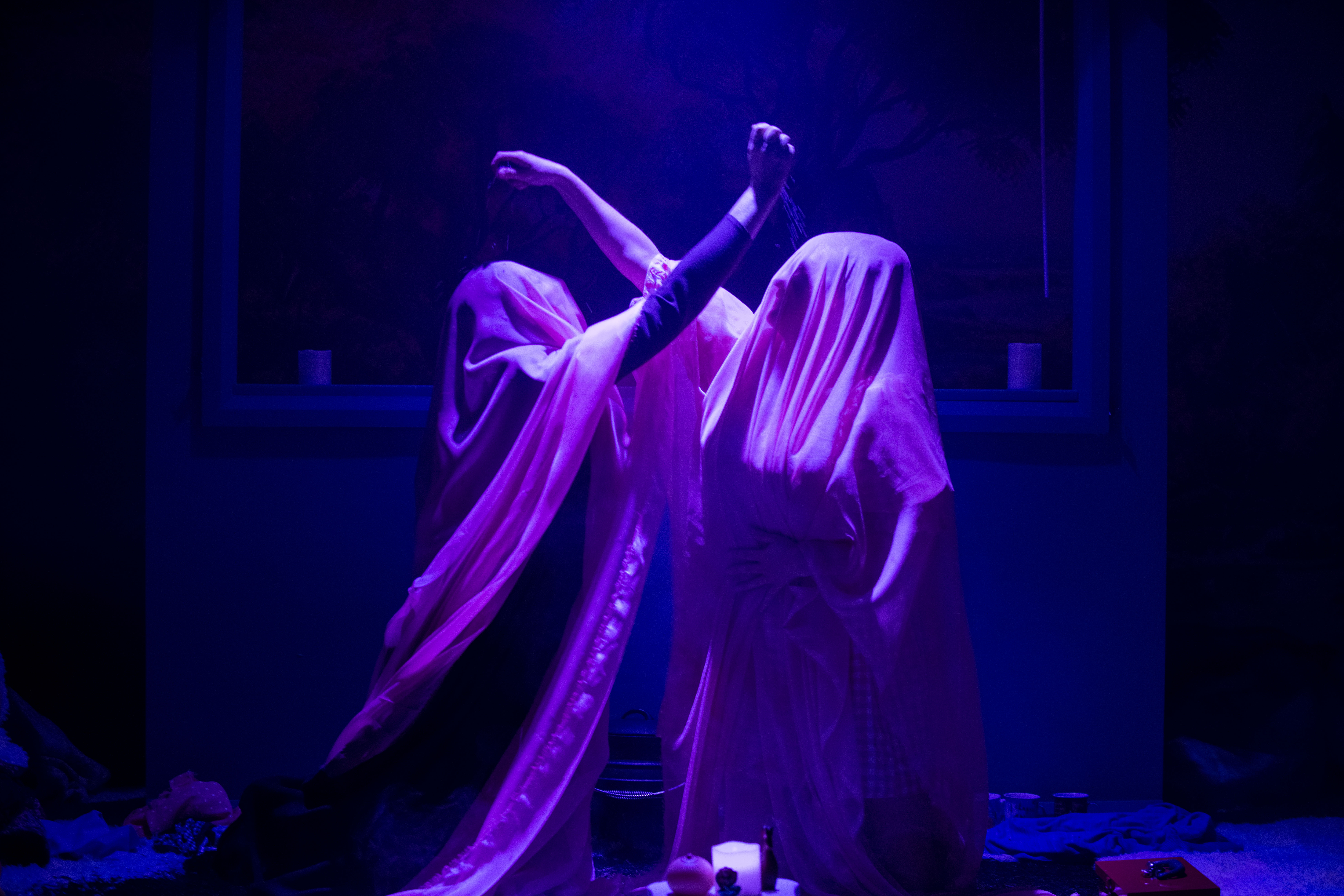 Two performers wearing veils have one arm extended above one another. They're illuminated by a purple spotlight.
