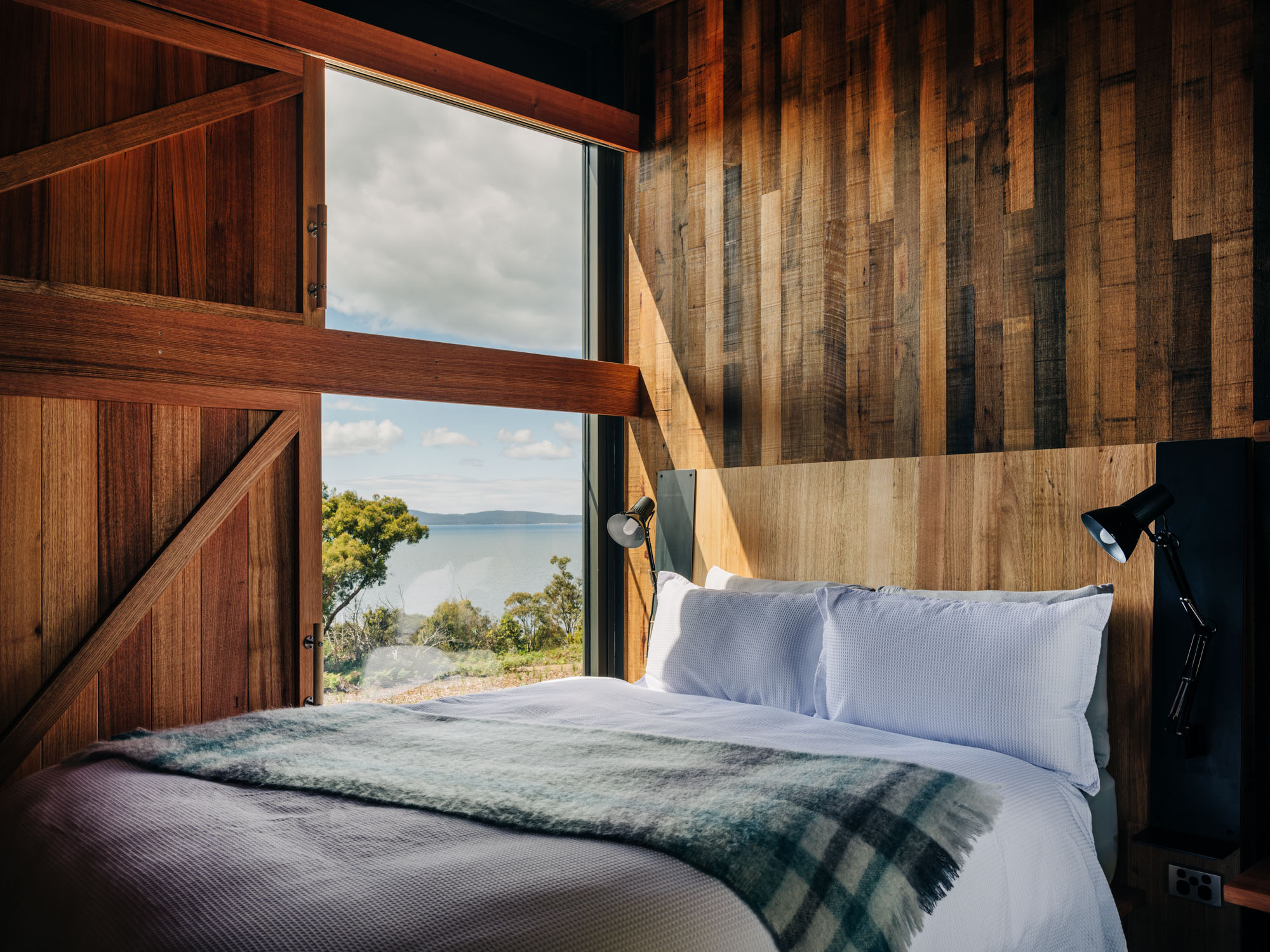 A bedroom with timber walls. From the bedside window, a view of the ocean.
