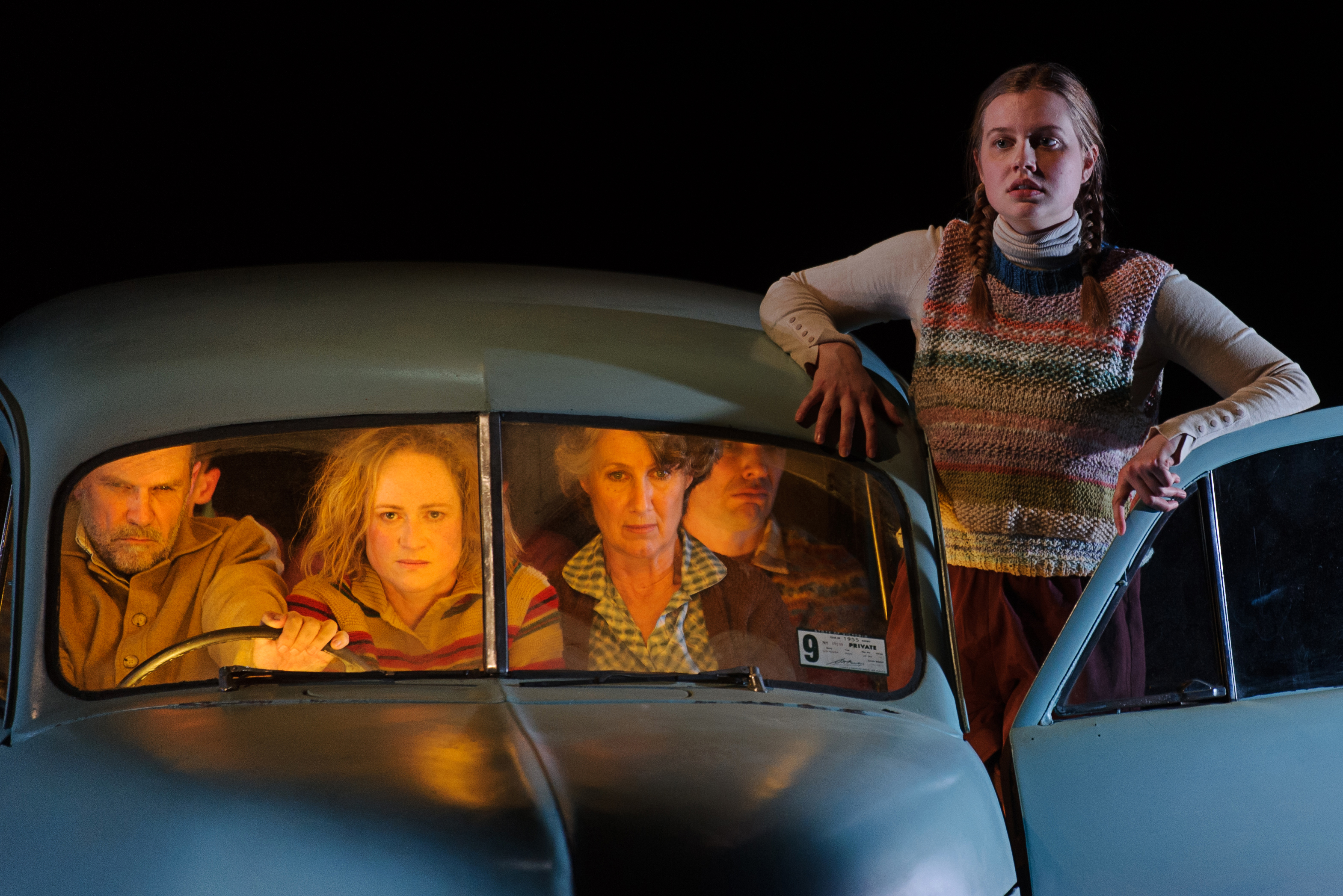 A family packed into a small car, while a young woman stands on the car's edge with the passenger door ajar.
