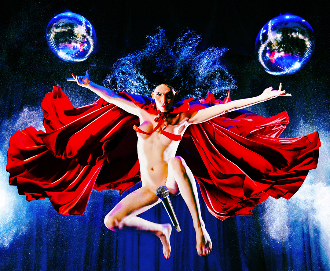 A naked performer with arms outstretched as they're suspended in the air. On either side of them is a disco ball, and covering their groin is a microphone.