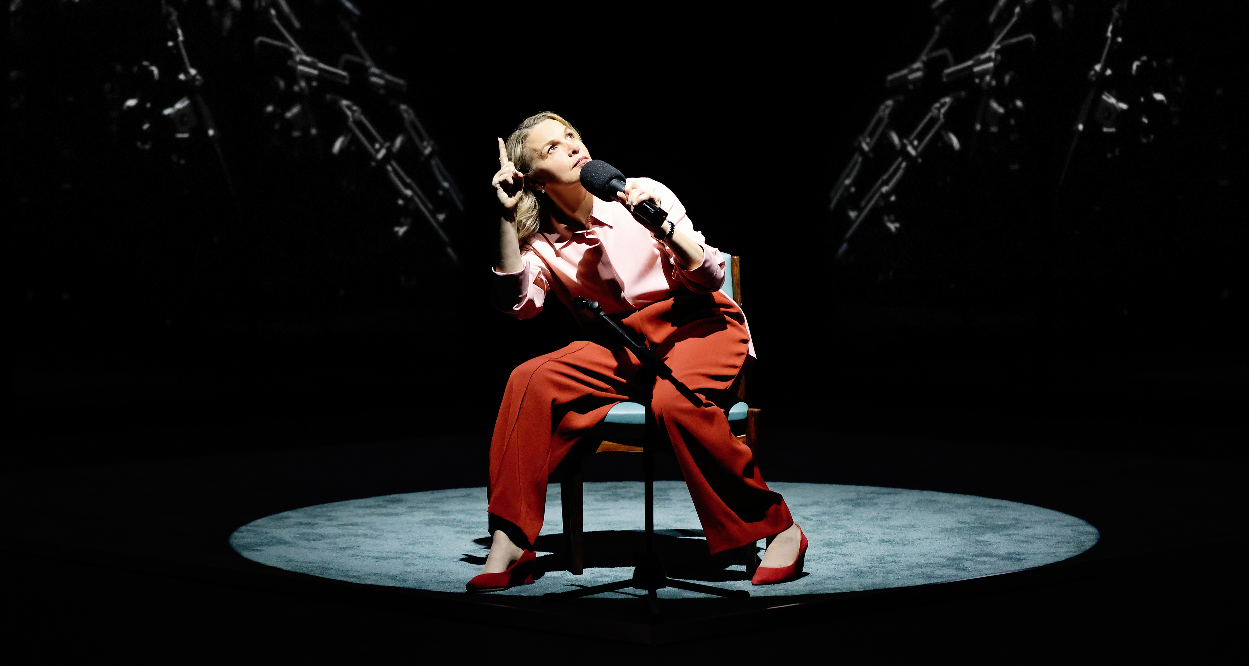Performer Justine Clarke sits under a spotlight on stage. She wears a red and pink outfit and holds a microphone.