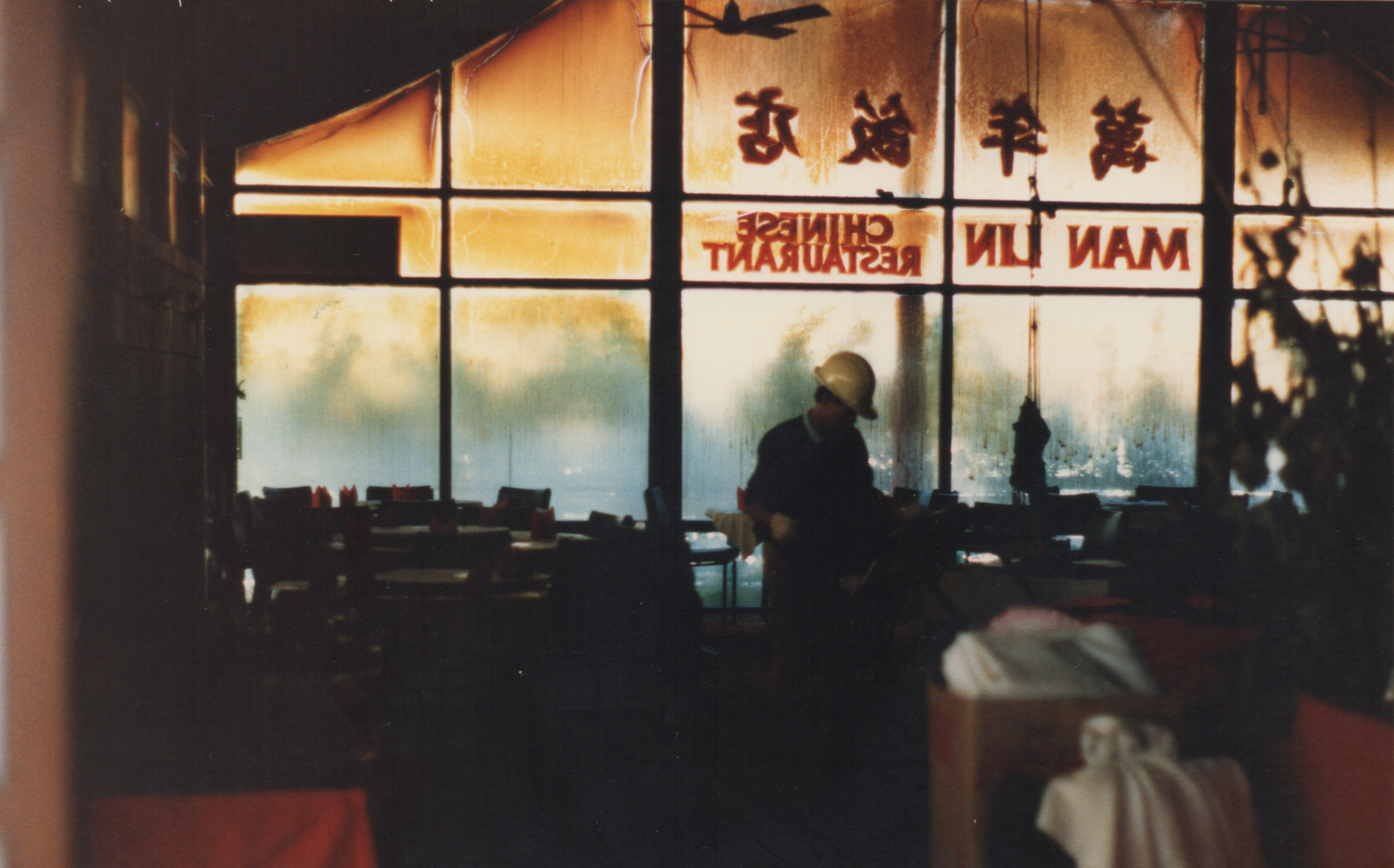 A man wearing a white helmet walks through a restaurant that has just suffered a firebomb attack.
