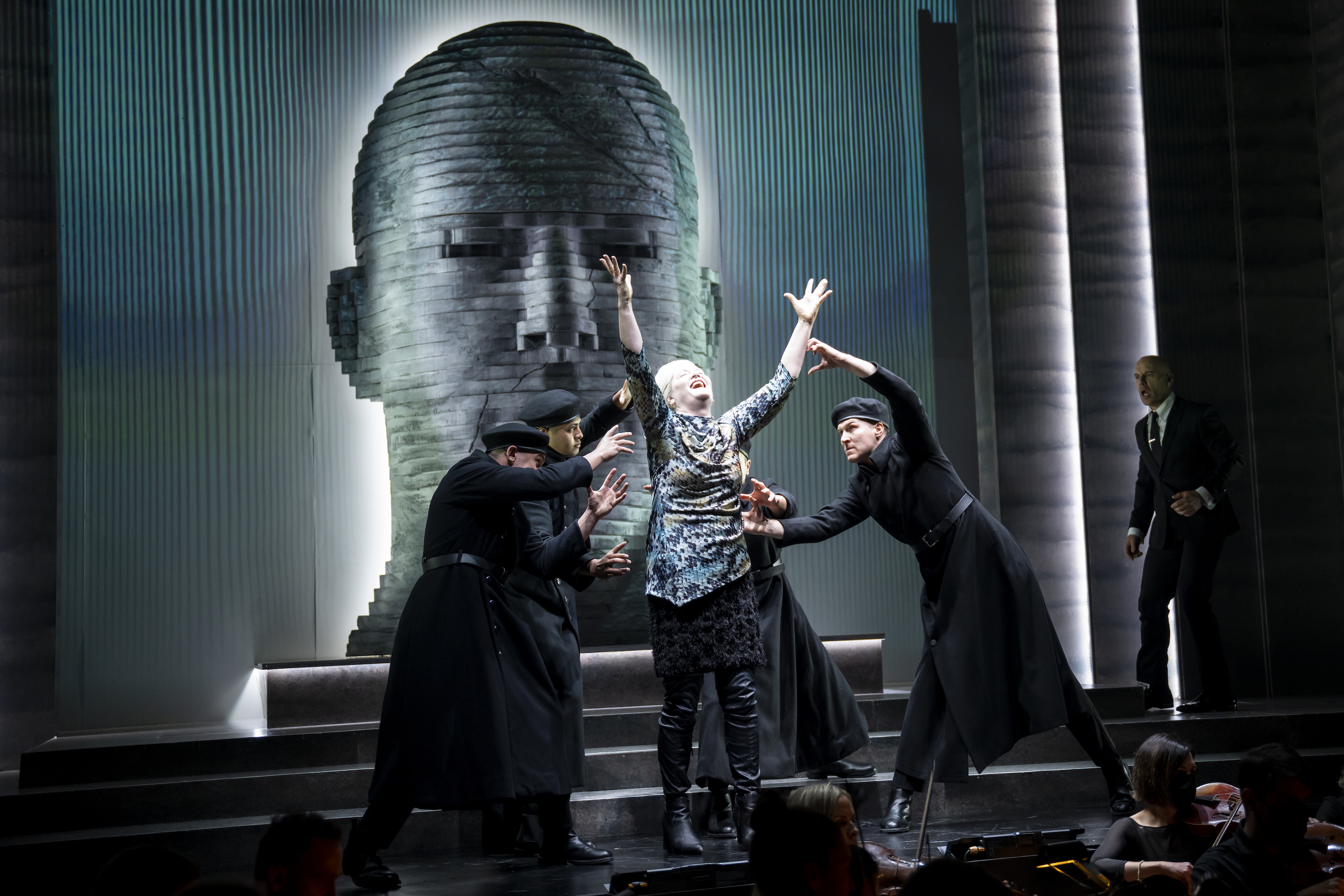 Four men in black clothing and berets surround a woman with white hair wearing a snake-patterned blue top on a dark stage. She has her arms outstretched above her and she appears to be singing. Behind the group is a screen showing a blue and grey distorted face made up on blocks that's somewhat unnerving. 