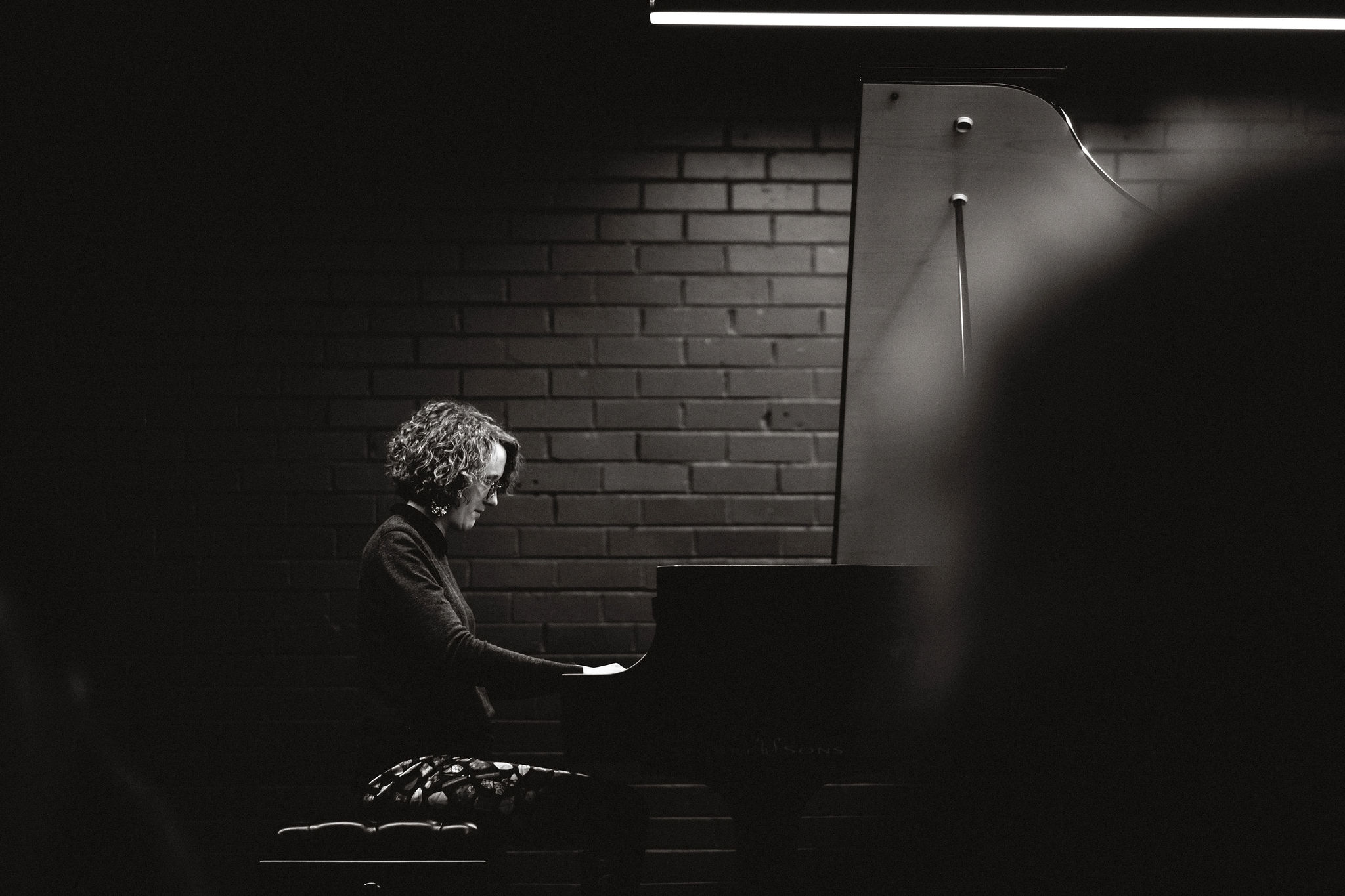 Black & white image of a woman with curly hair sitting at a piano, her eyes watching her hands on the keys. To her side and in the background is a brick wall.
