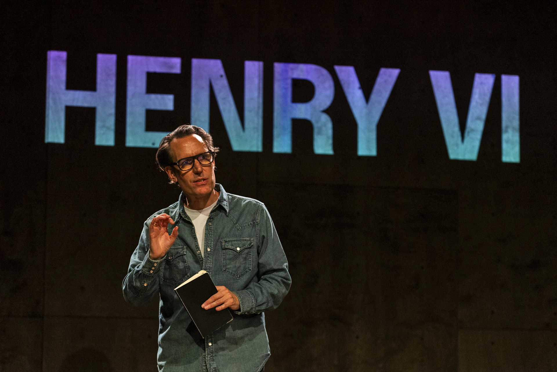 Peter Evans on stage with a sign reading "Henry VI" behind him. 