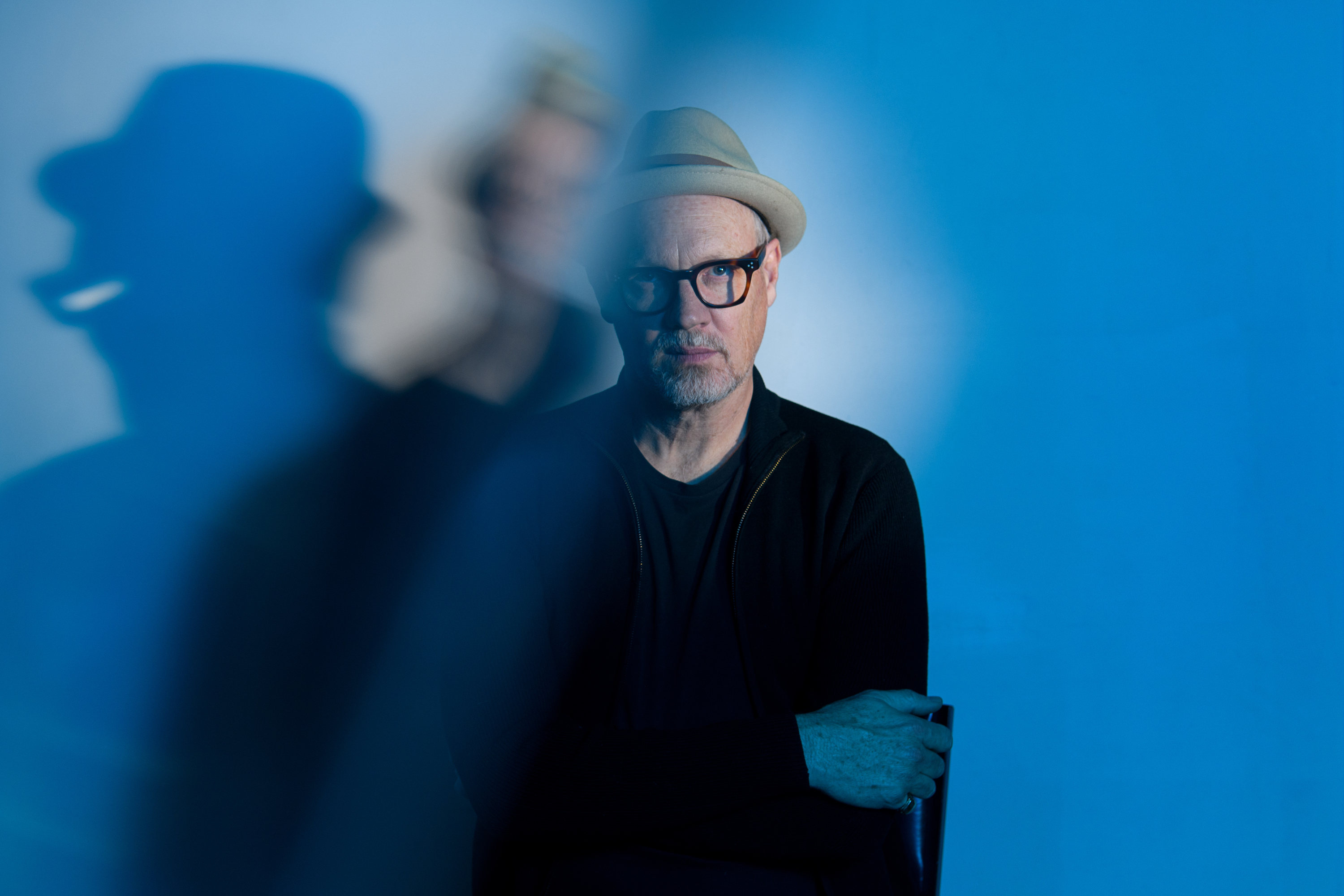 A Caucasian man wearing a beige fedora hat, tortoiseshell glasses and dark clothing stands in front of a blue background, with the left side of the image slightly blurred and mirrored and his shadow apparent on the wall