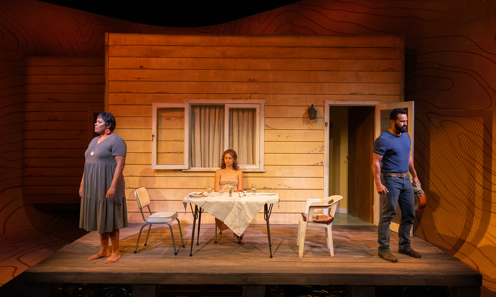Three performers on stage. One sits at a table in the centre, while the others stand on either side of the stage, faced away from each other.