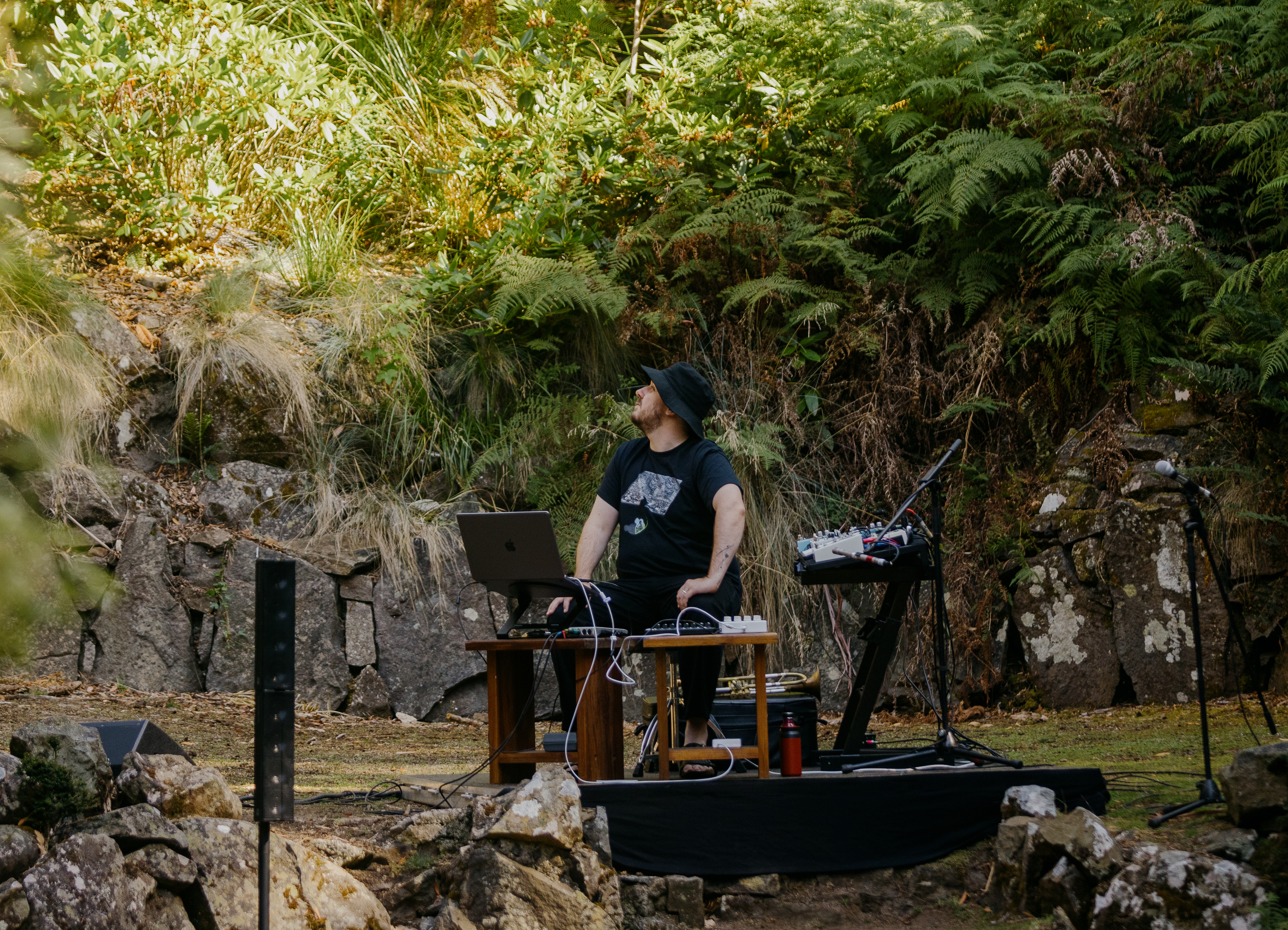 Performer Reuben Lewis sits outside, on a small stage with his electronic equipment. He is looking up at what surrounds him – moss-covered rock and bushland bathed in sunlight.