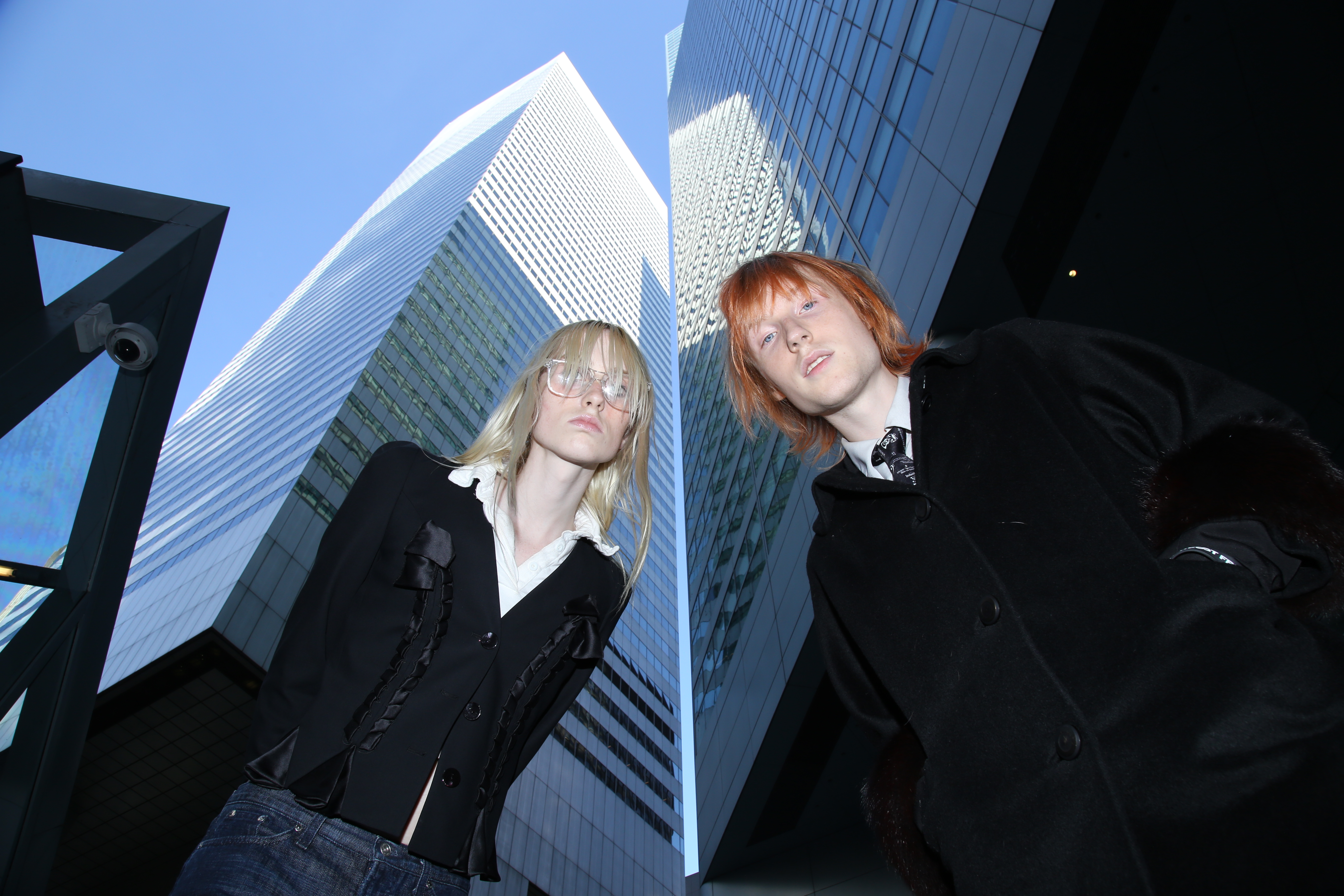 Two people with long hair standing underneath skyscrapers.