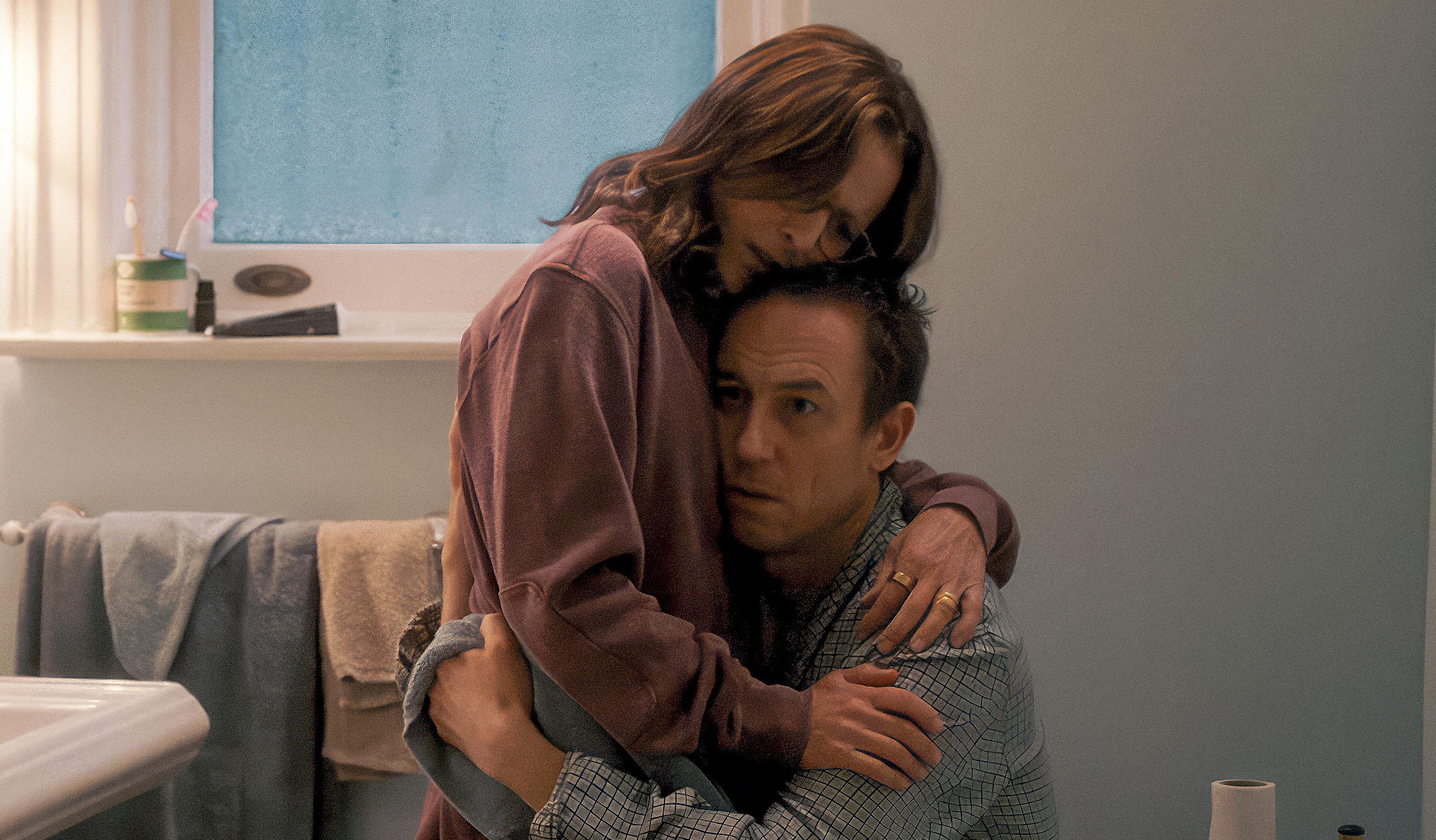 A woman stands in a bathroom holding a man's face against her chest.