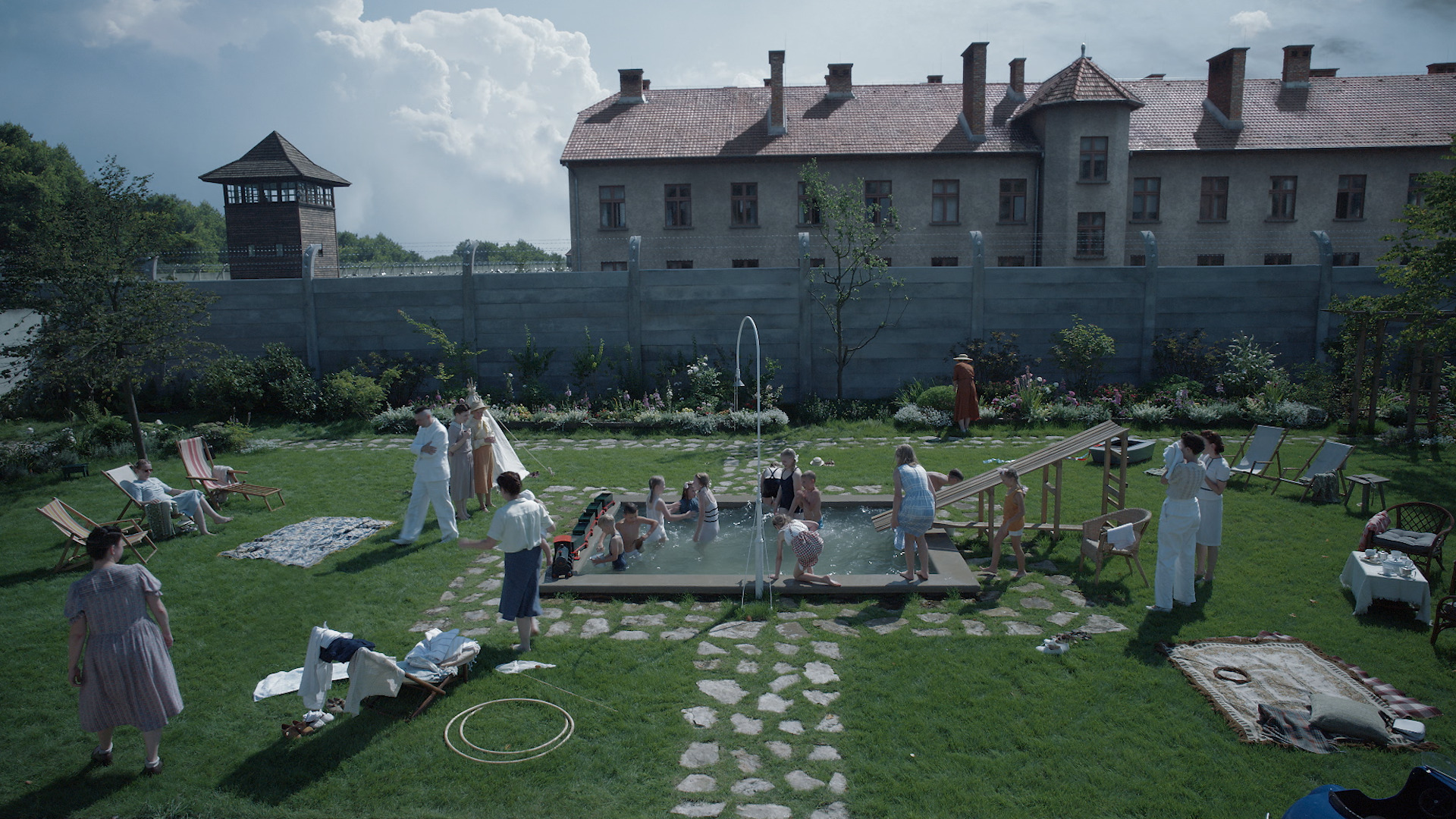 Children play in a backyard pool. On the other side of the fence is the building of a concentration camp.
