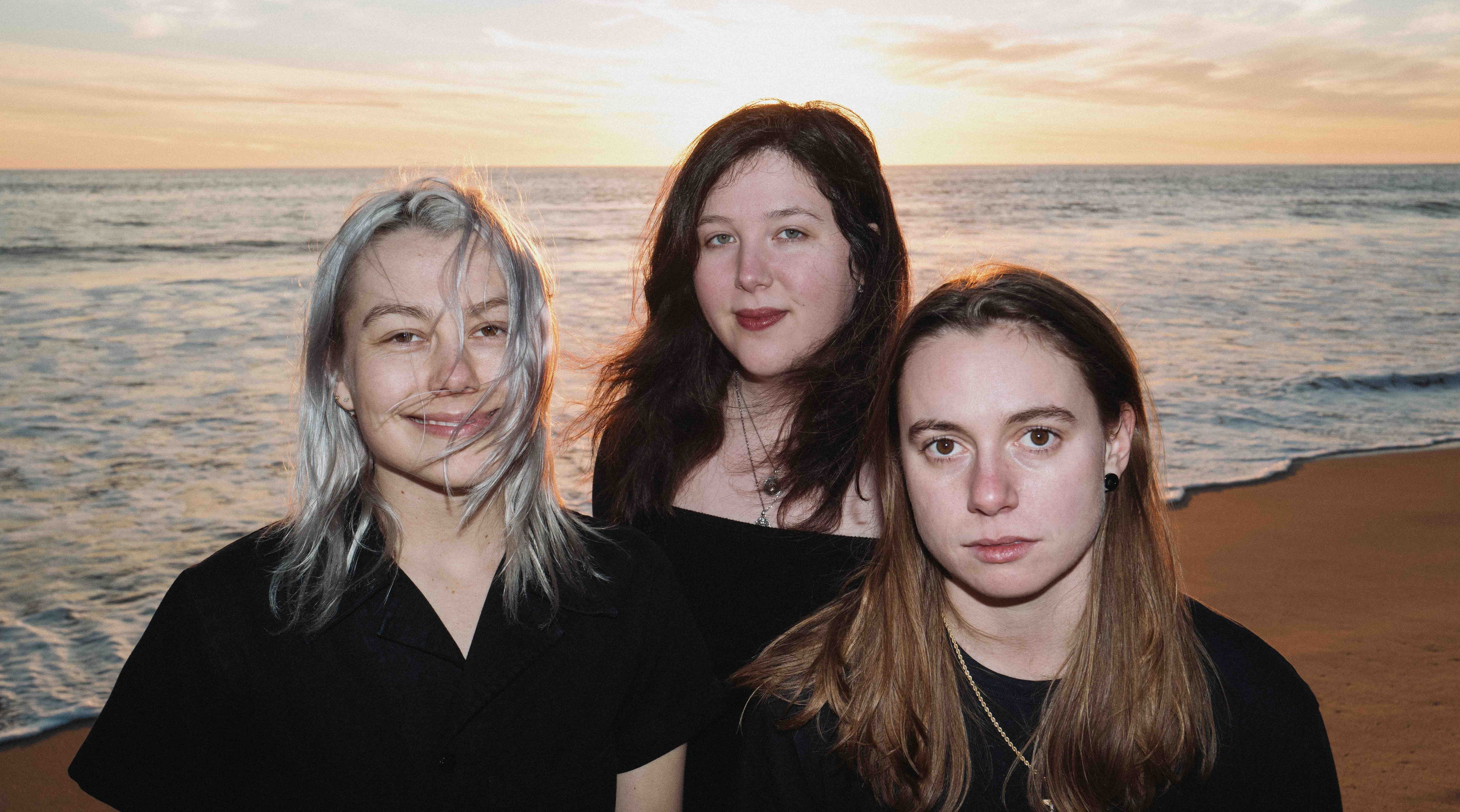 Members of boygenius standing on the shoreline of a beach with the sun setting behind them.