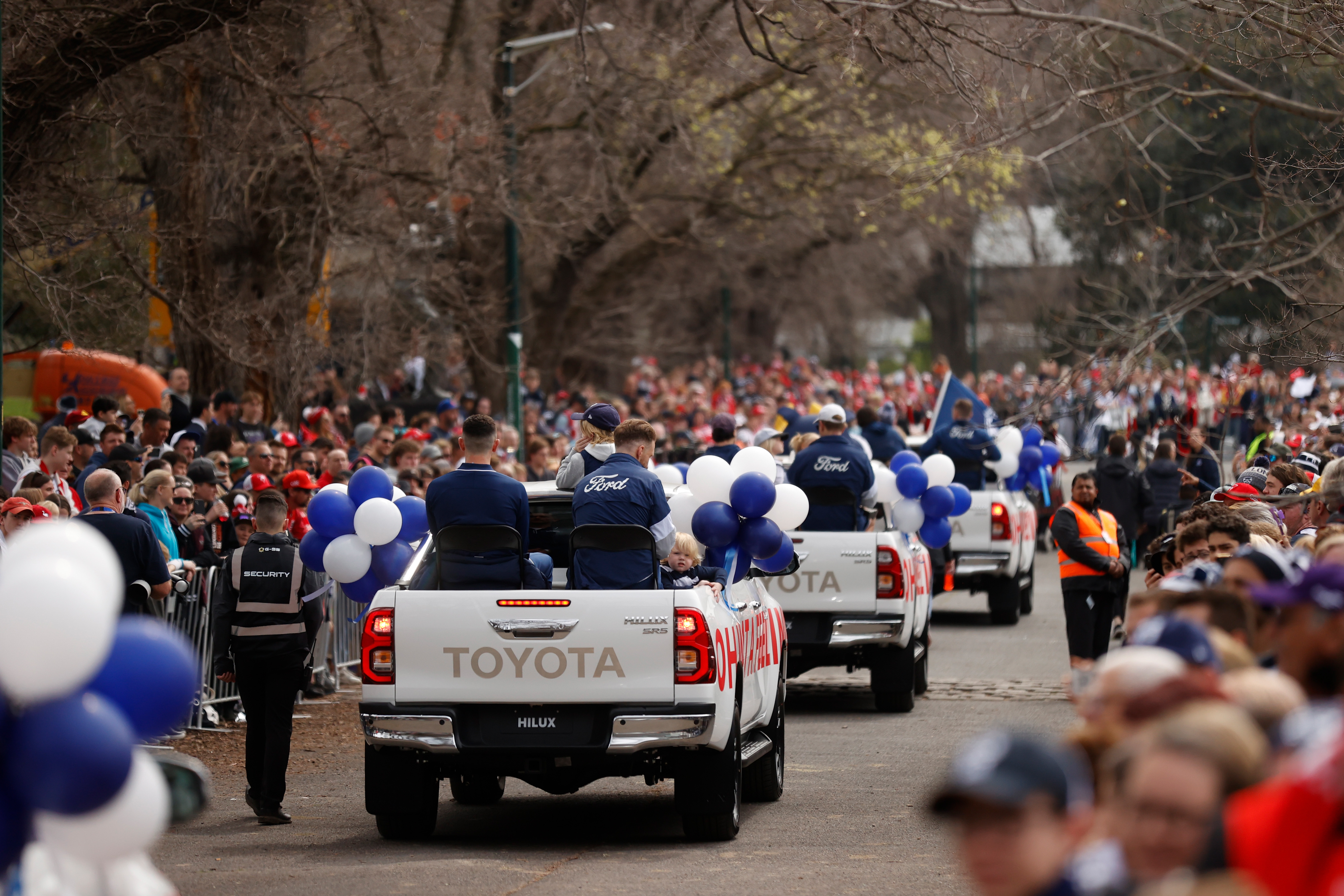 Geelong Cats players ride in Toyota Hilux’s while fans watch behind a fence.