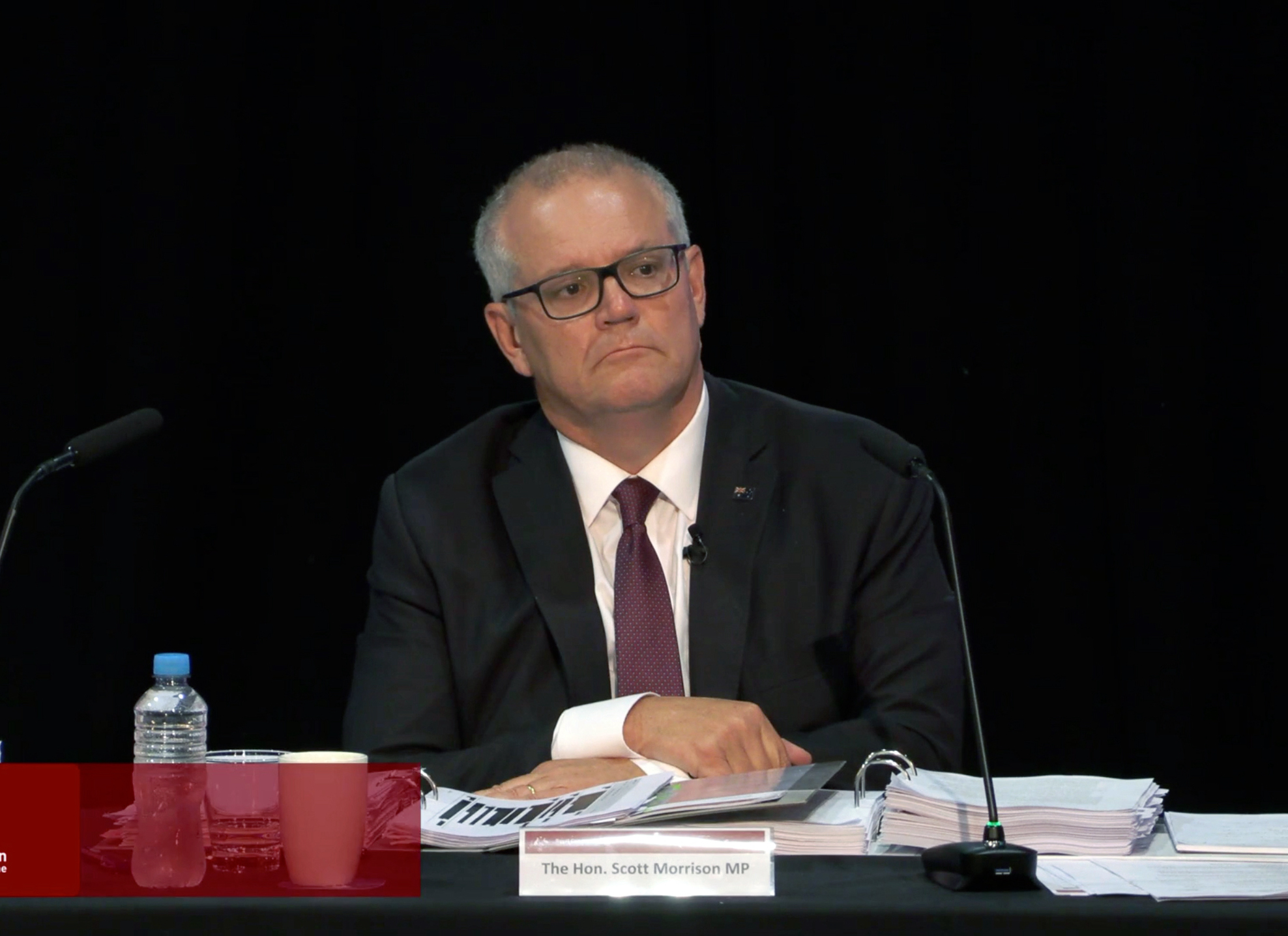 A balding, middle-aged Caucasian man wearing rectangular black glasses, a dark suit and a boring red tie sits in front of a desk loaded with files and papers, and two microphones. He looks bored, and there is a paper stand with "The Hon. Scott Morrison MP" in front of him.