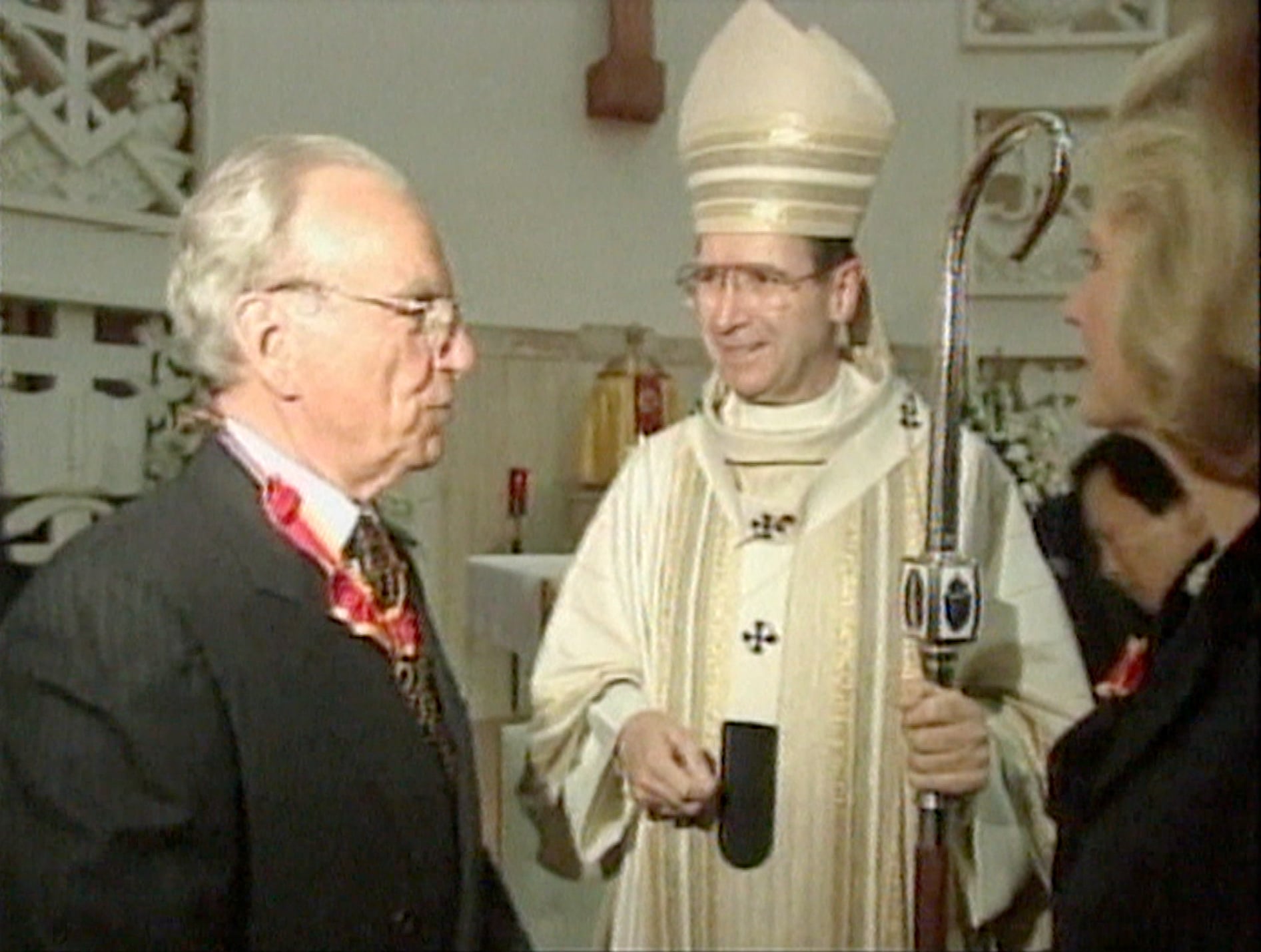 An older Caucasian man in a suit is wearing some sort of red decoration around his neck, while a Catholic bishop in a white robe, tall hat and holding a cane looks on. The picture is grainy and obviously from the late 90s.