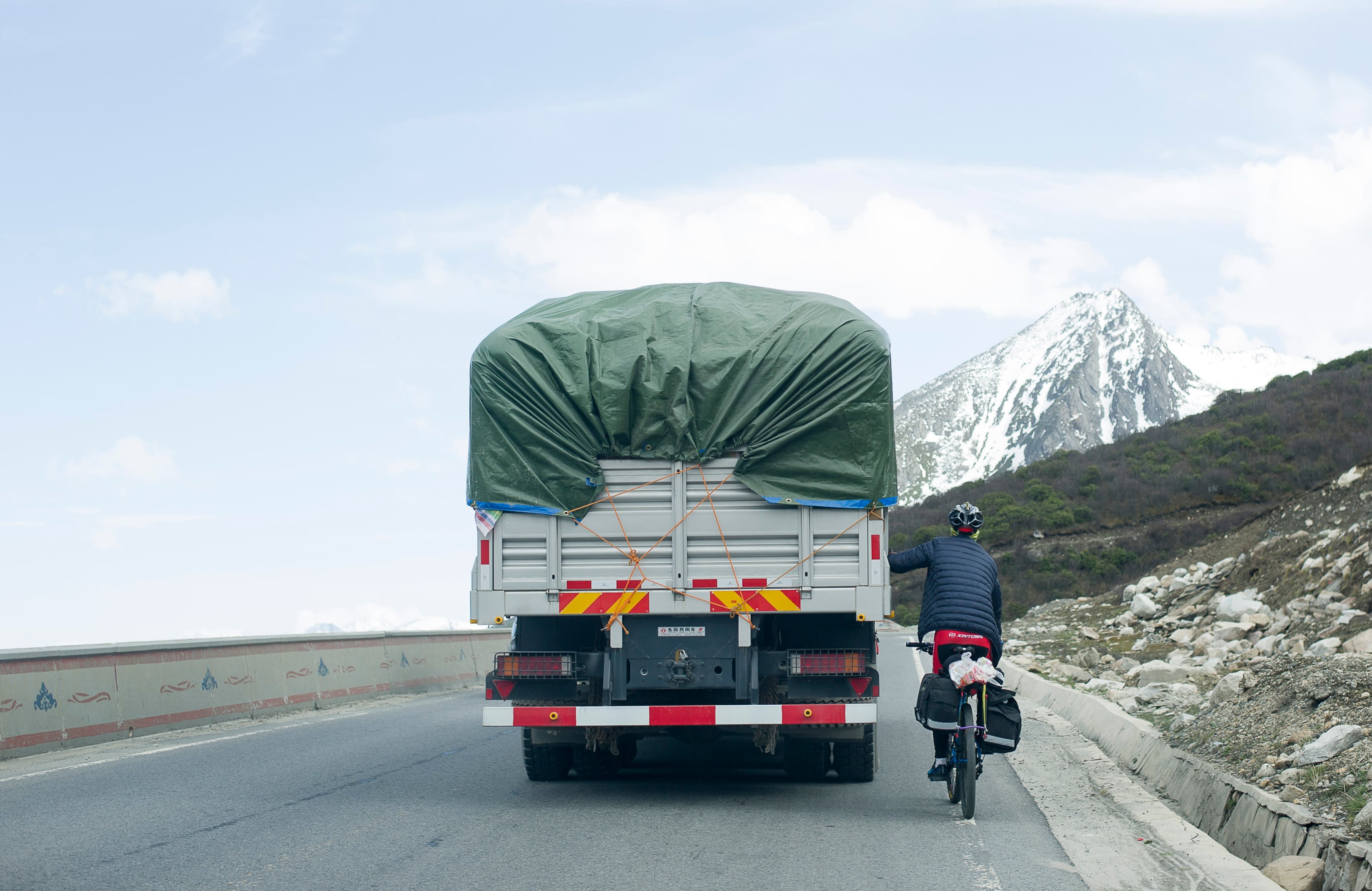 A cyclist hitches a ride with a truck on the Sichuan–Tibet Highway, with a snow mountain looming in the background.