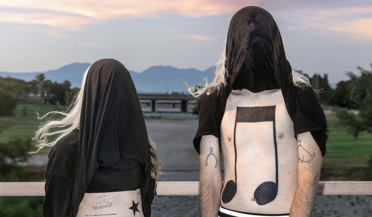 Album cover: two figures standing on a bridge, the bottom of their black t-shirts pulled over their faces. Their exposed bodies reveal an array of tattoos, including a large musical note