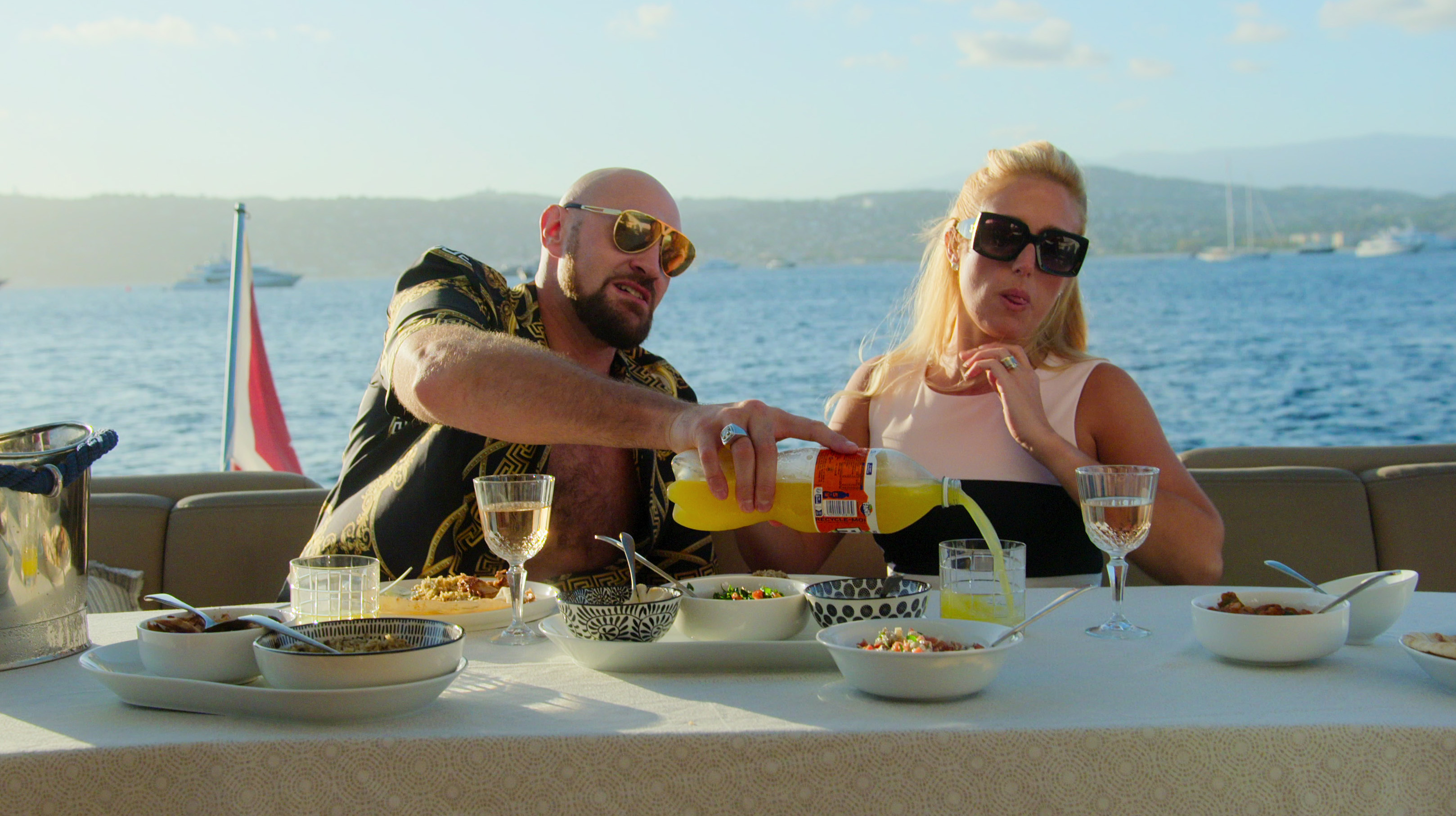 A couple wearing sunglasses sit at a table piled with food and drinks by the water. 