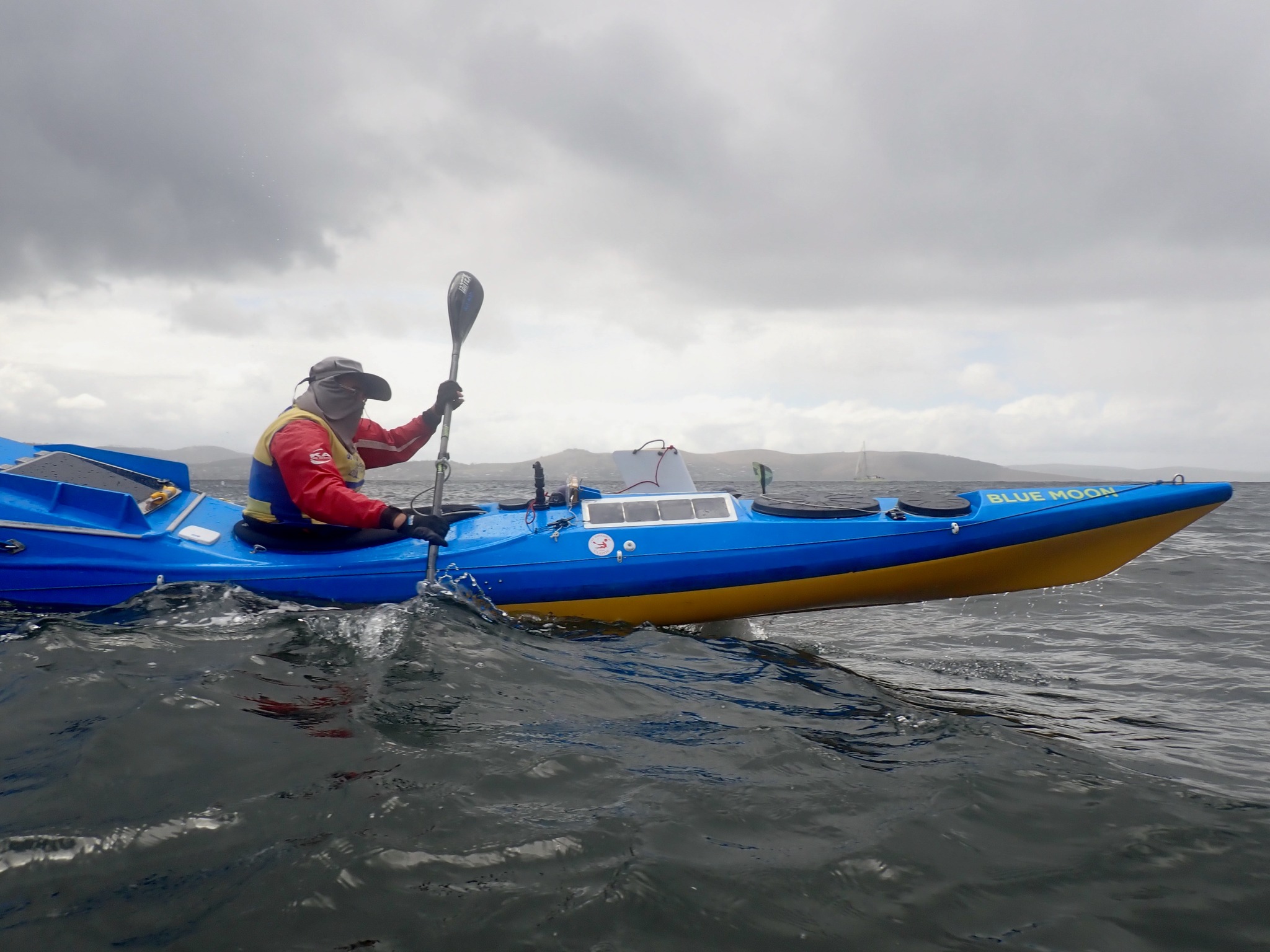 A man in weather/water protection gear paddling a large blue kayak with "Blue Moon" written on its side. The ocean is grey and the sky cloudy, with land distant on the horizon.