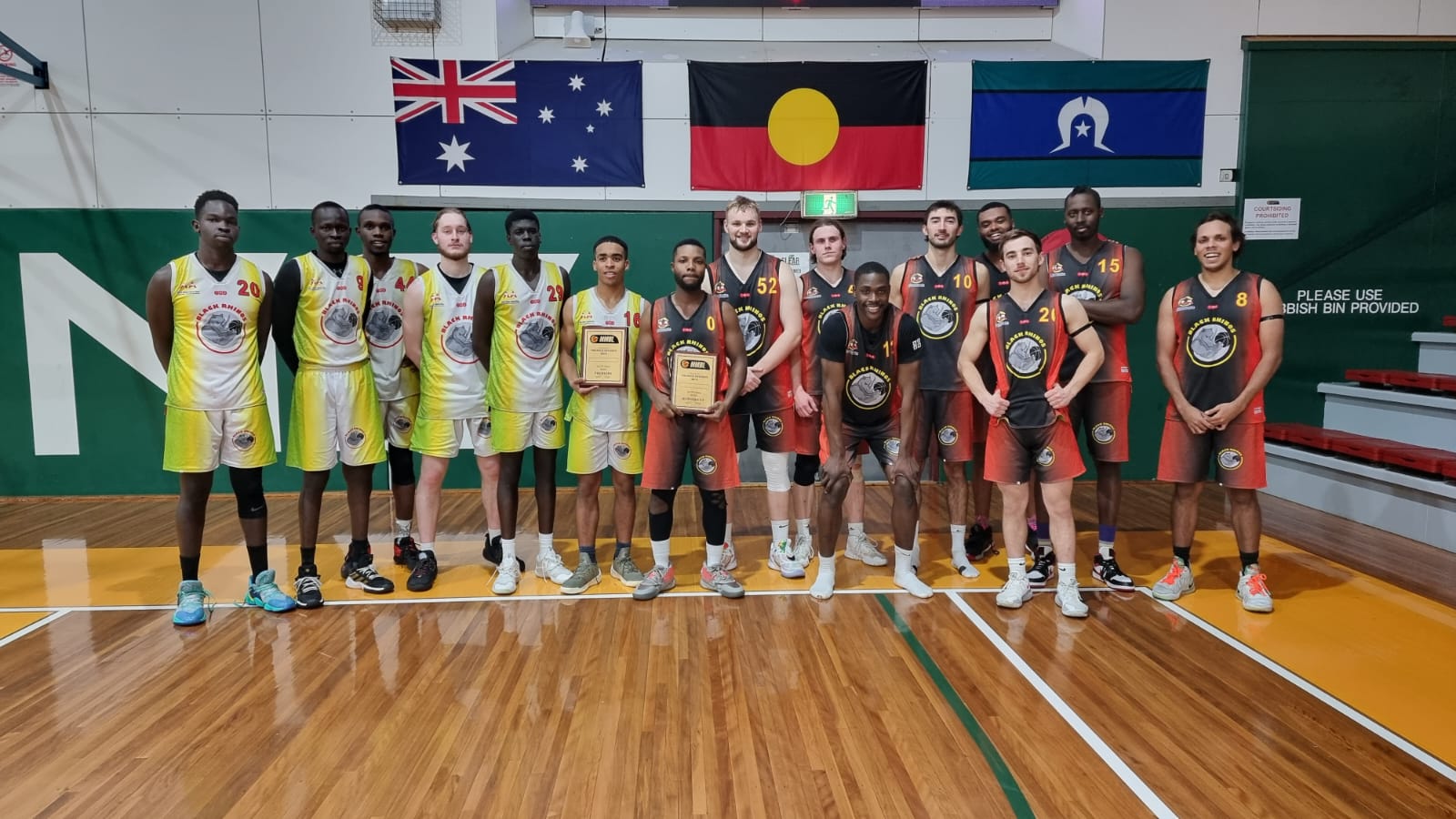 The Black Rhinos basketball team stand on the edge of a basketball court with their awards, underneath national flags.