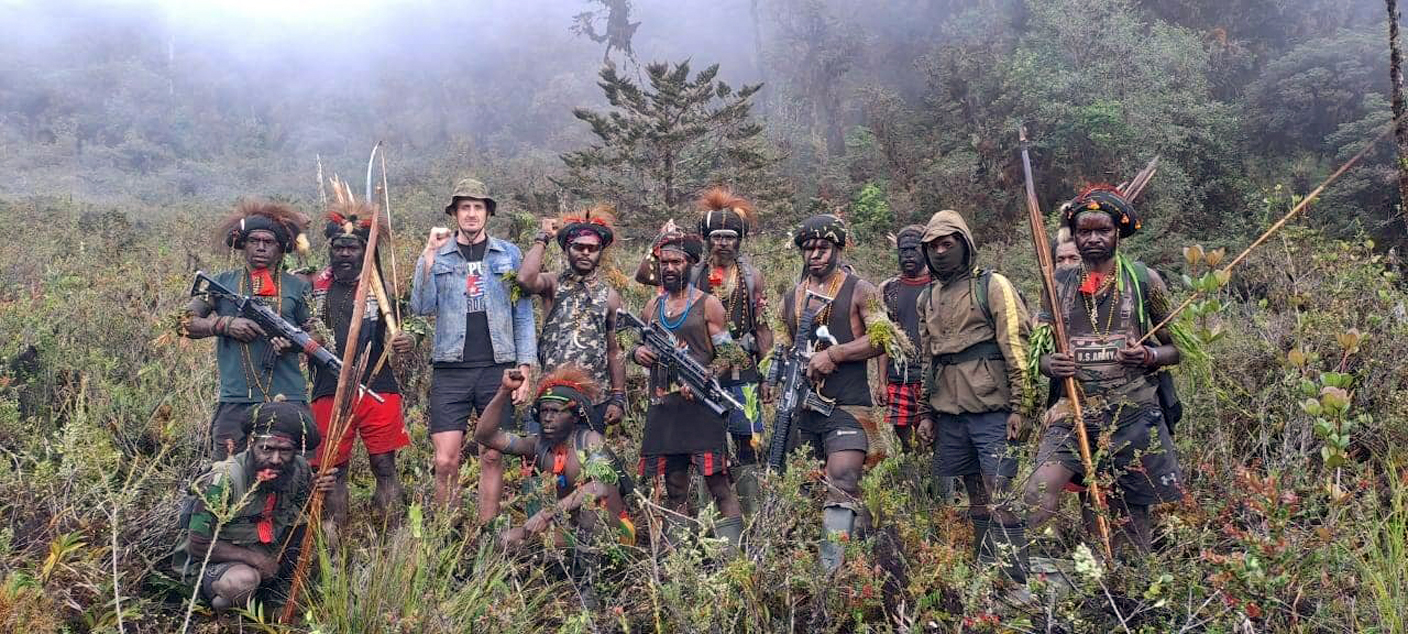 Phillip Mark Mehrtens wearing a blue denim jacket and standing with his fist raised. He is flanked on both sides with his separatists – Papua separatists who hold machine guns.