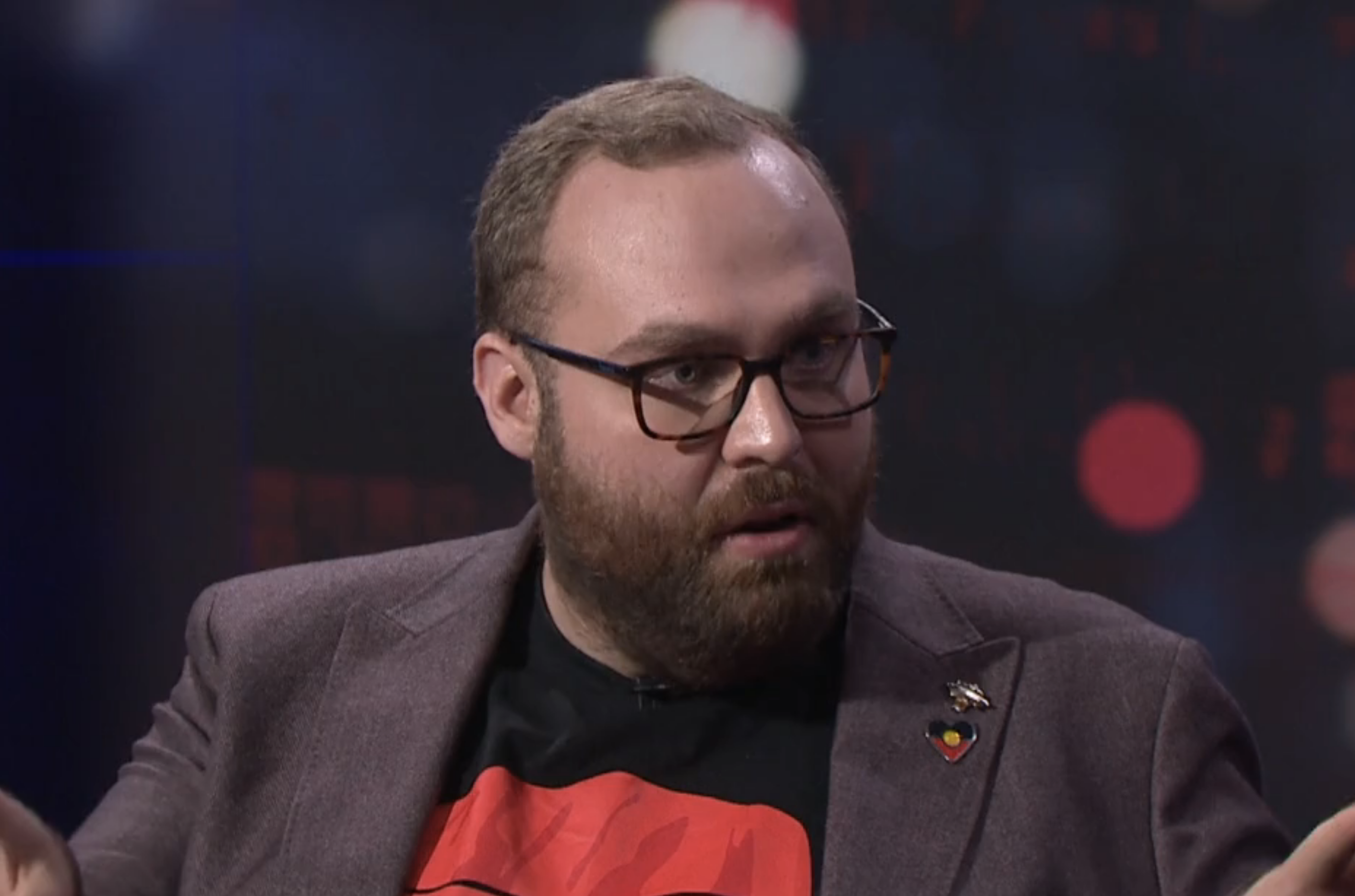 A man wearing glasses and a badge of the Australian Aboriginal flag speaks on a panel.