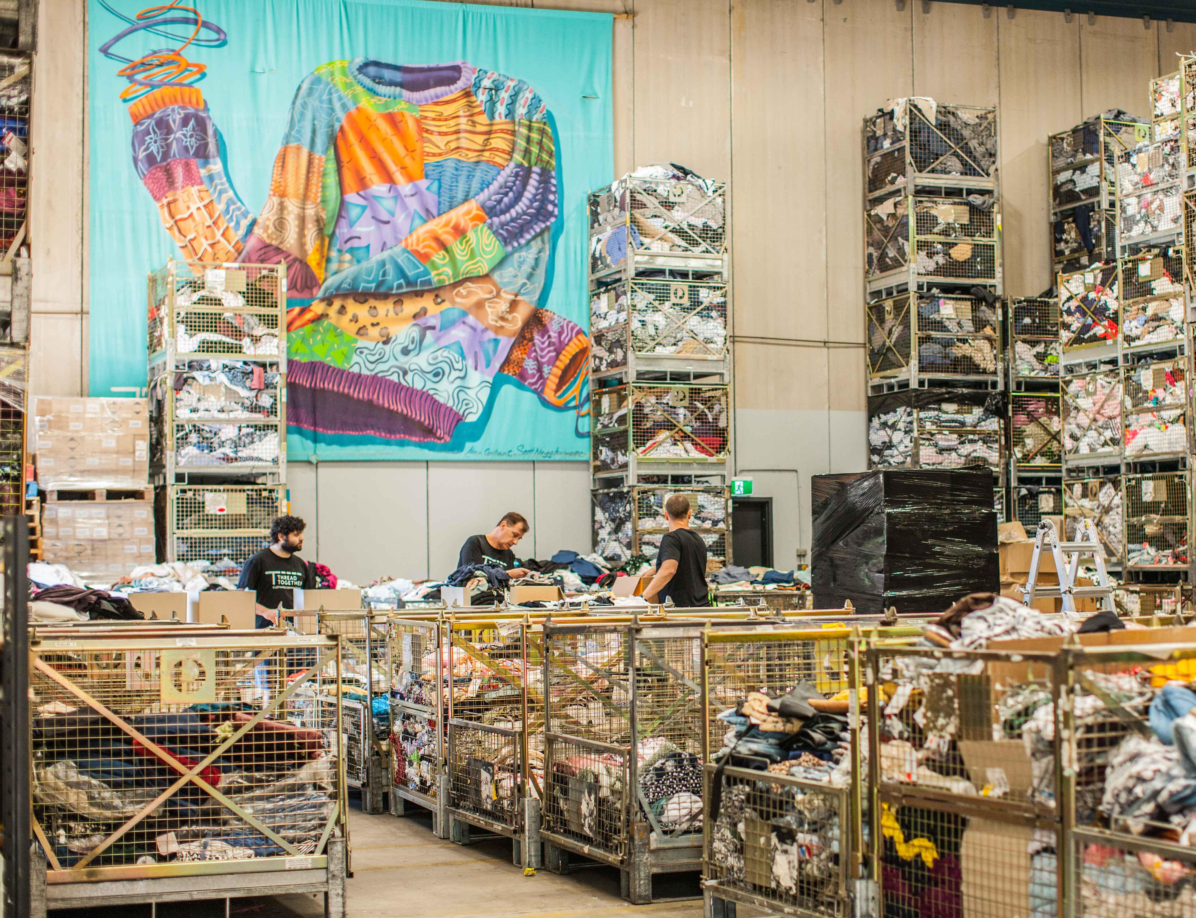 A warehouse filled with clothes that are separated into metal cages. A a large banner with the print of a colourful knit hangs from the wall as workers sort clothes.