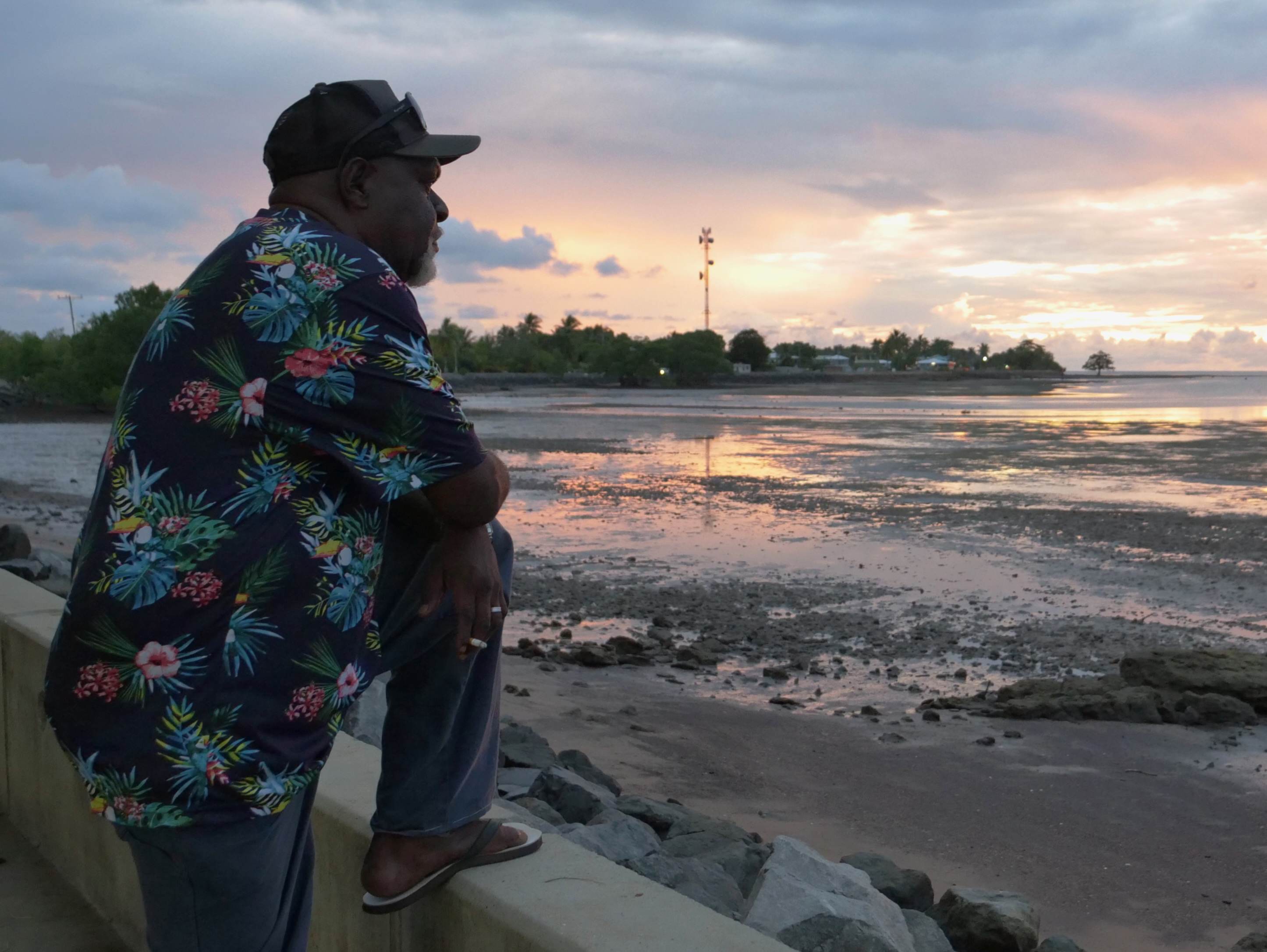 A man wearing a floral shirt and a cap gazes beyond the coastline under a sunset.