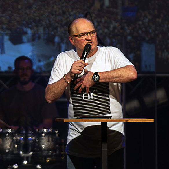 A man wearing glasses and a t-shirt speaking on stage with a microphone in his hand.