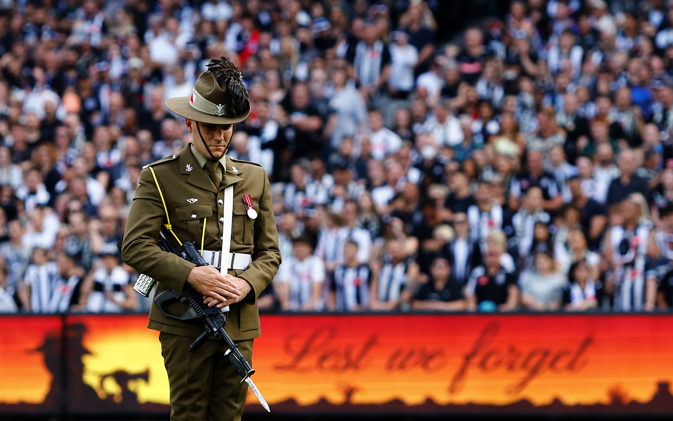 A soldier stands in uniform and with a rifle on his hip, head bowed in the middle of a football field with fans in striped jerseys in the stands.