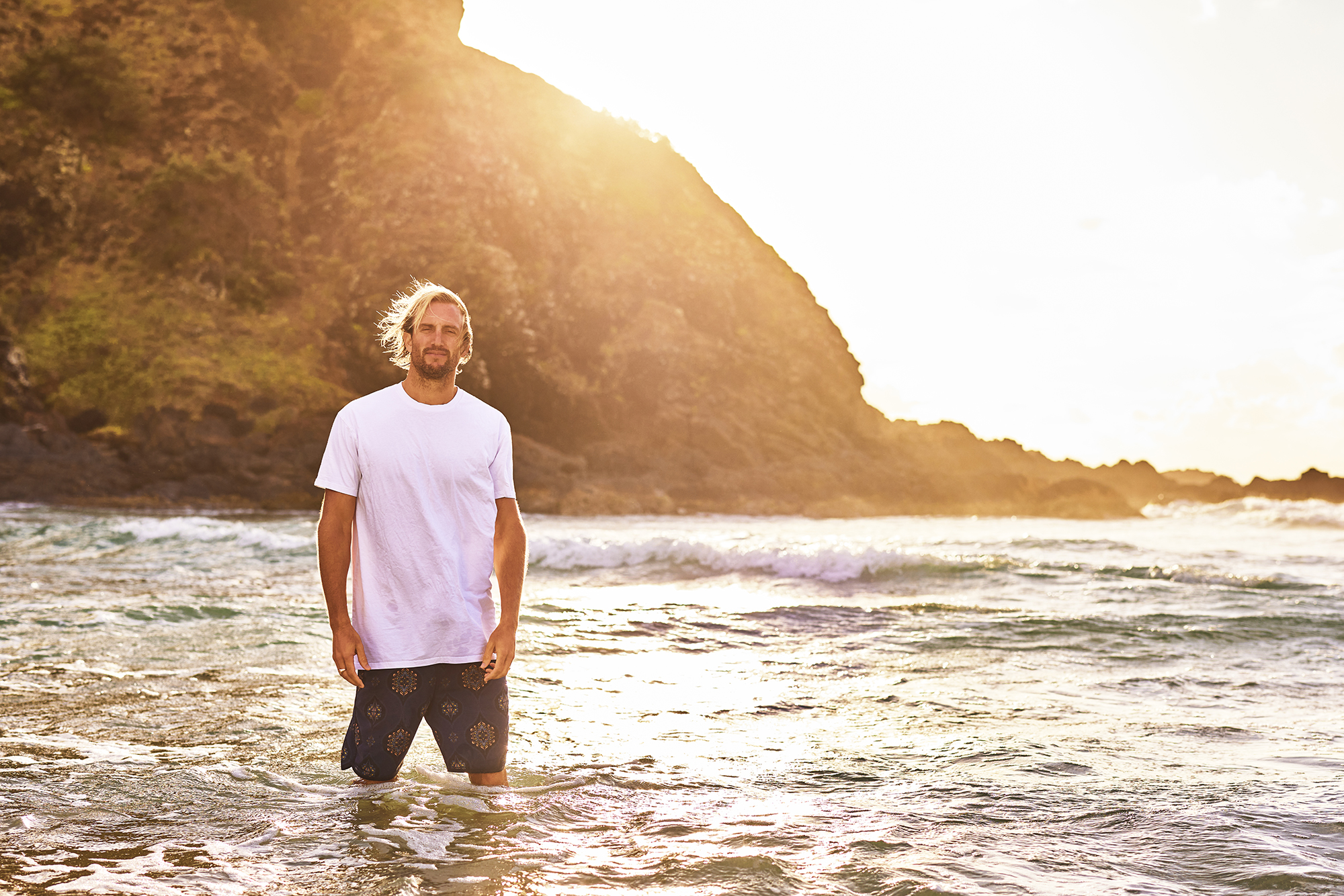 A tall man with long hair and wearing a white t-shirt in stands in knee-high water. The sun sets behind the coastline.