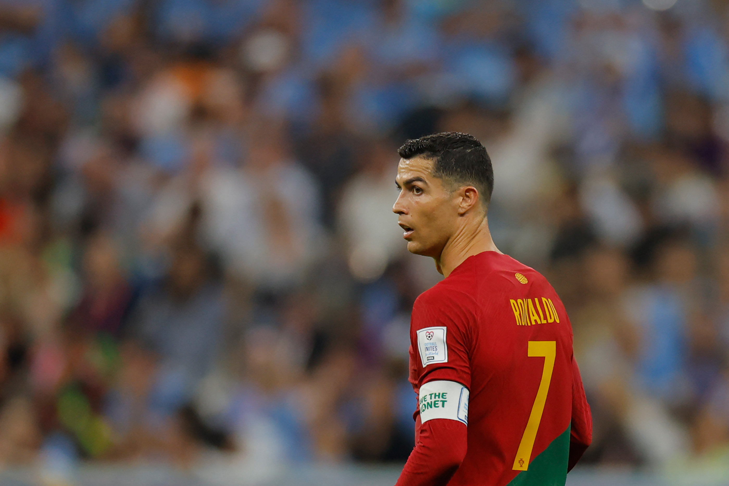 A side profile shot of the soccer player Cristiano Ronaldo; he has cropped brown hair, is wearing the red and yellow Portuguese football uniform and is looking to the left of the photo. A blurred crowd behind him shows that he is on the soccer pitch at a game.