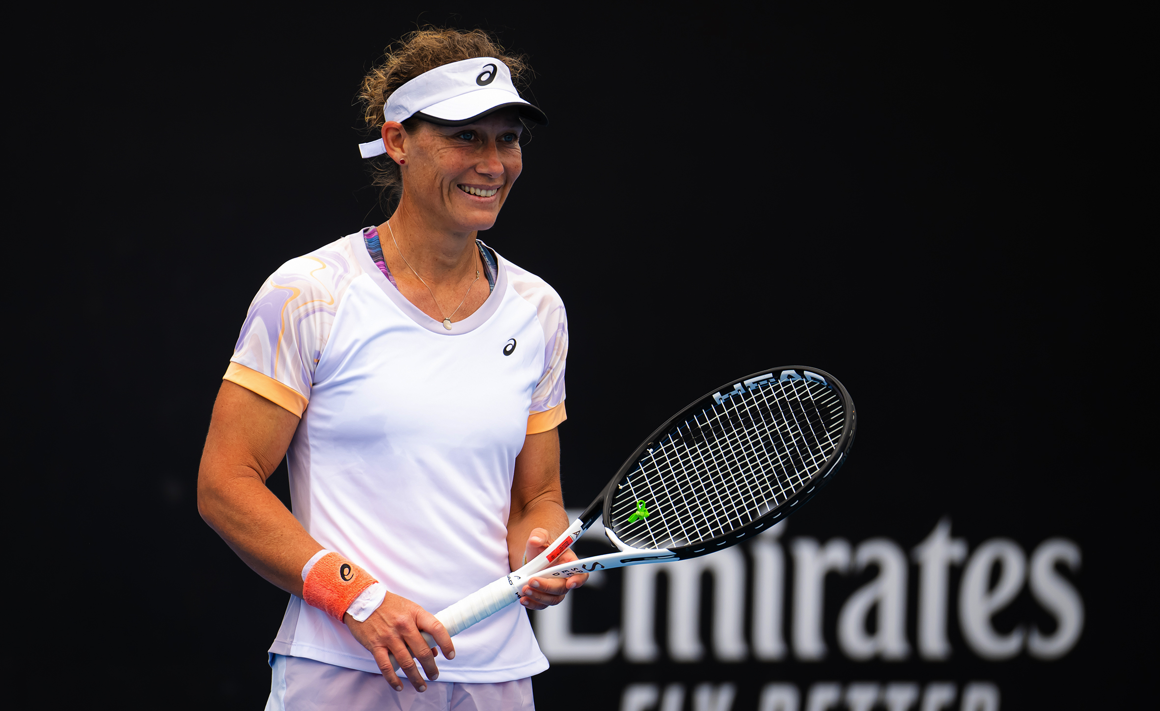 A Caucasian woman in her thirties appears in white tennis gear holding a tennis racket and wearing a visor. She is smiling