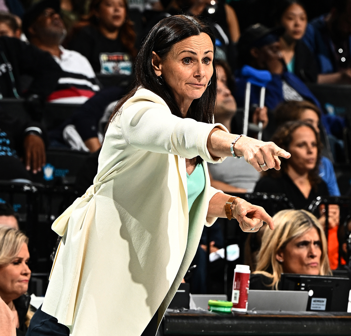 A coach with dark hair directs players on a basketball court. A crowd watches in the stands behind her.