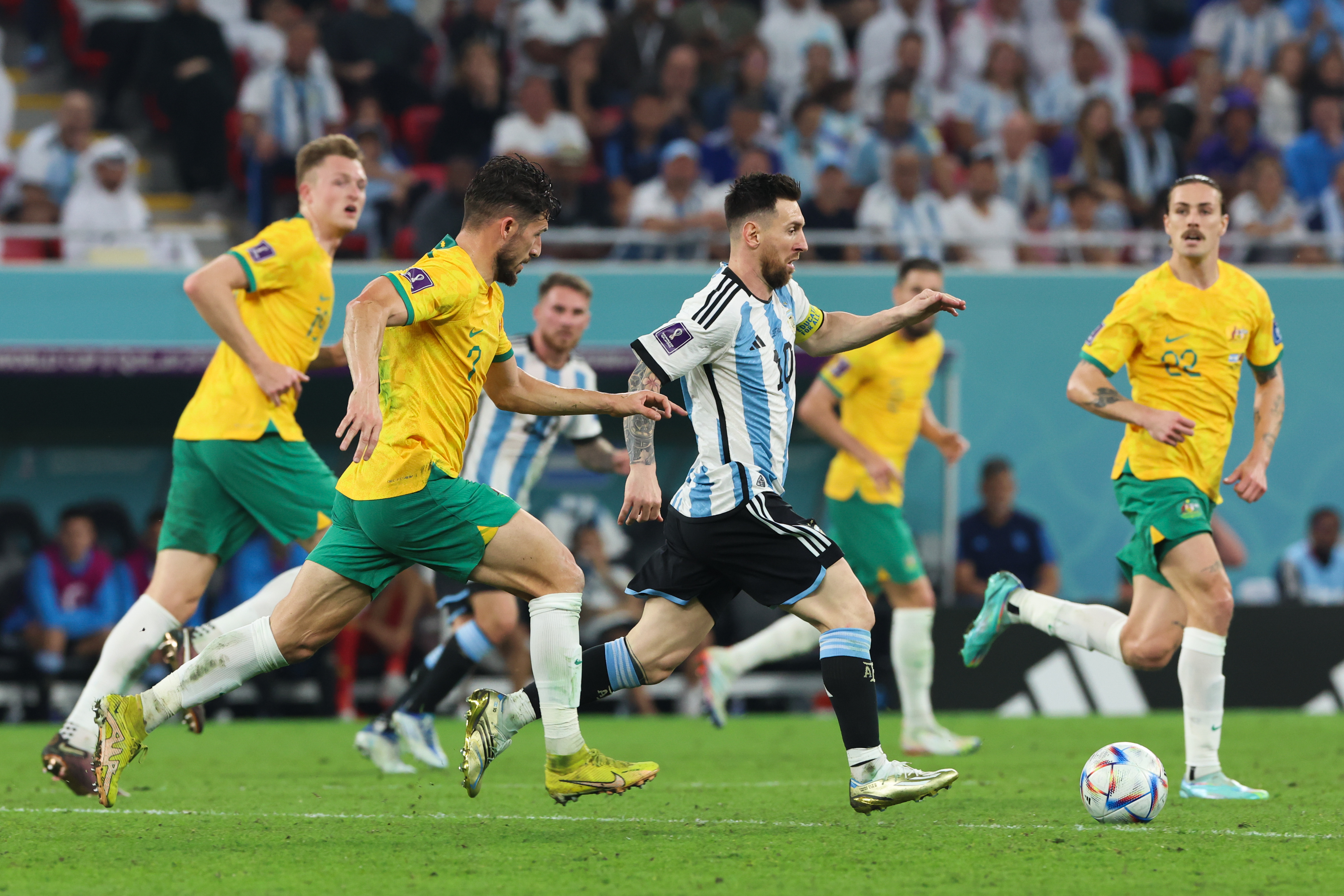 Four men on a soccer pitch are in the foreground of the image; three are wearing the green shorts and gold shirts of the Australian soccer team, and the fourth man in the centre is wearing black shorts and a blue and white striped shirt (the Argentinian team's uniform). This man is Lionel Messi, and he has possession of a soccer ball. 