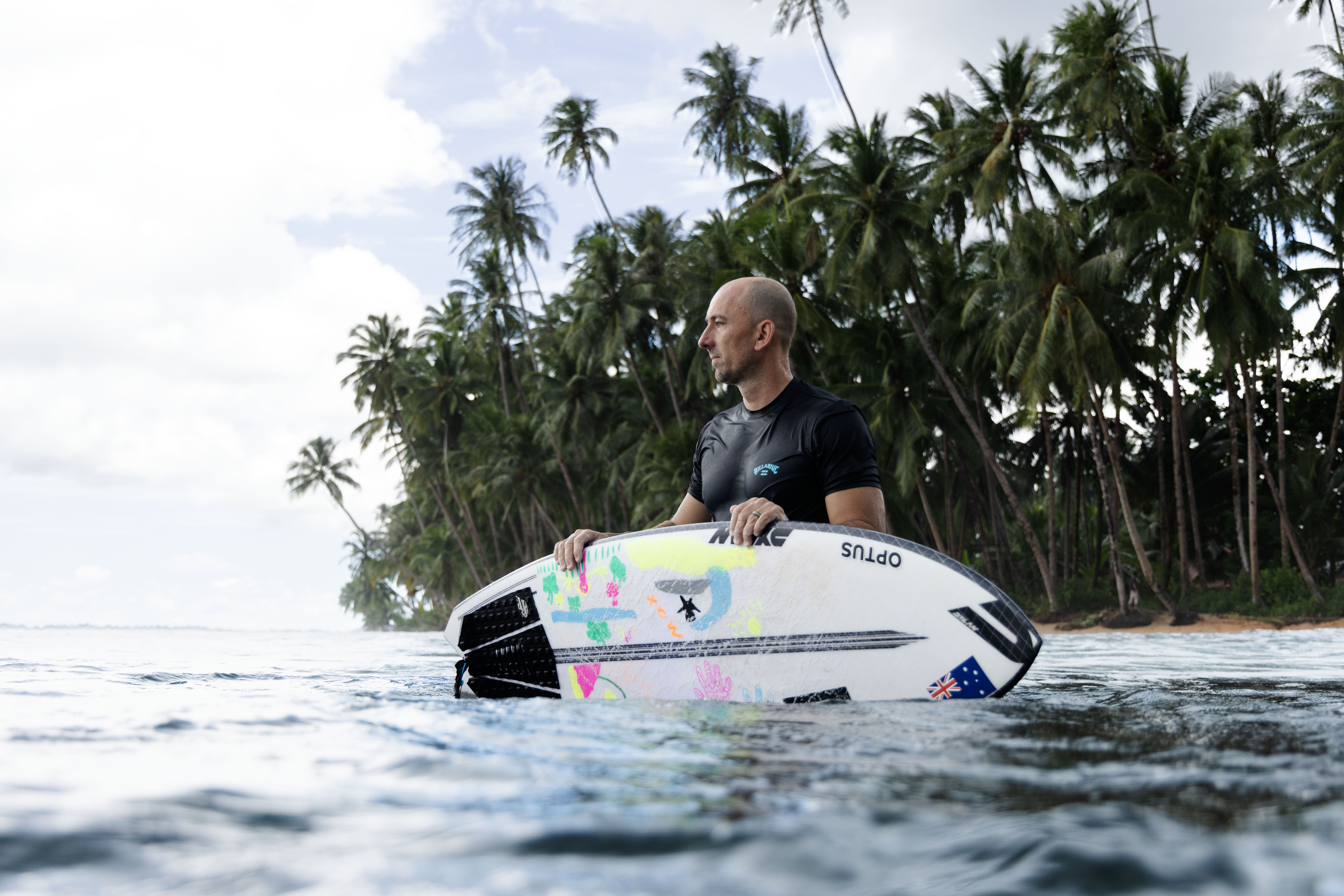 A surfer stands in the shallows with his surfboard floating in the water.