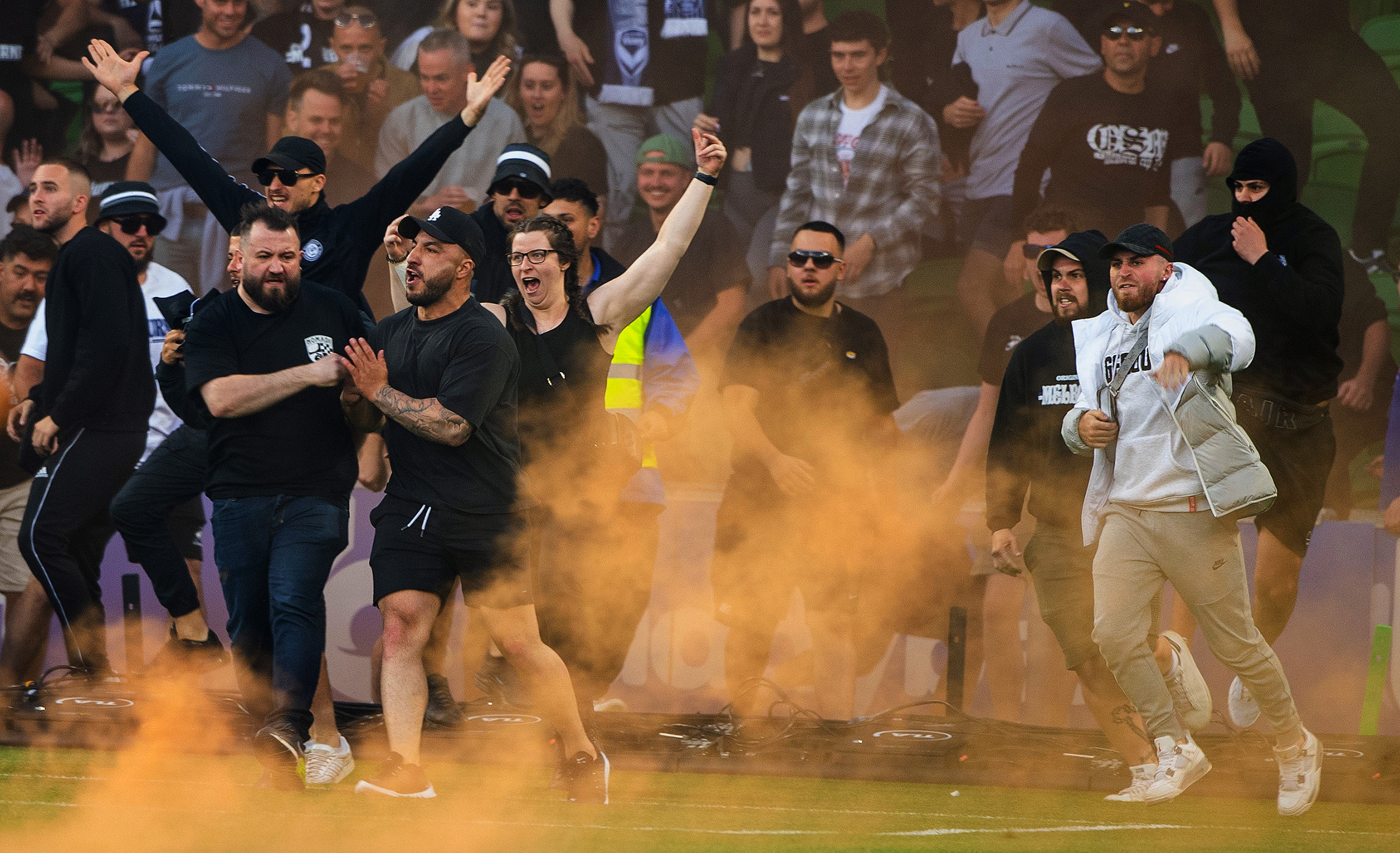 People wearing black clothing, sunglasses and some in hoodies appear to be storming onto a football pitch looking angry and yellow, while yellow smoke from a flare swirls around them