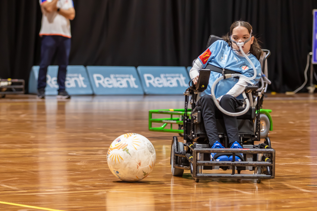 A powerchair football player on court with the ball beside her.