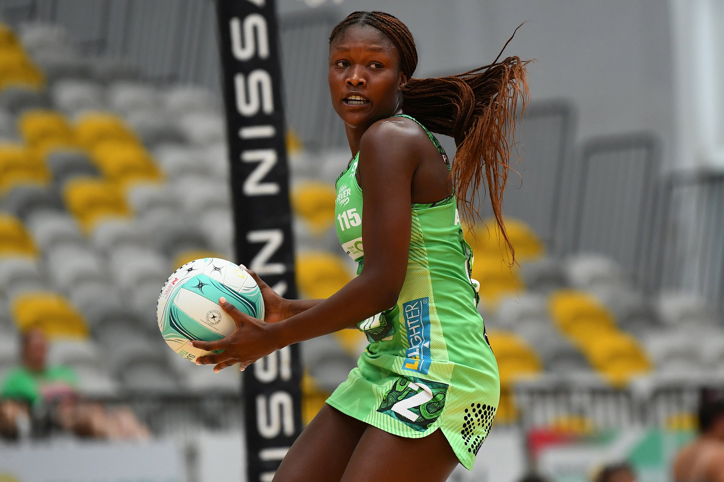 A netball player on court with the ball in her hands, and empty stand behind her. 