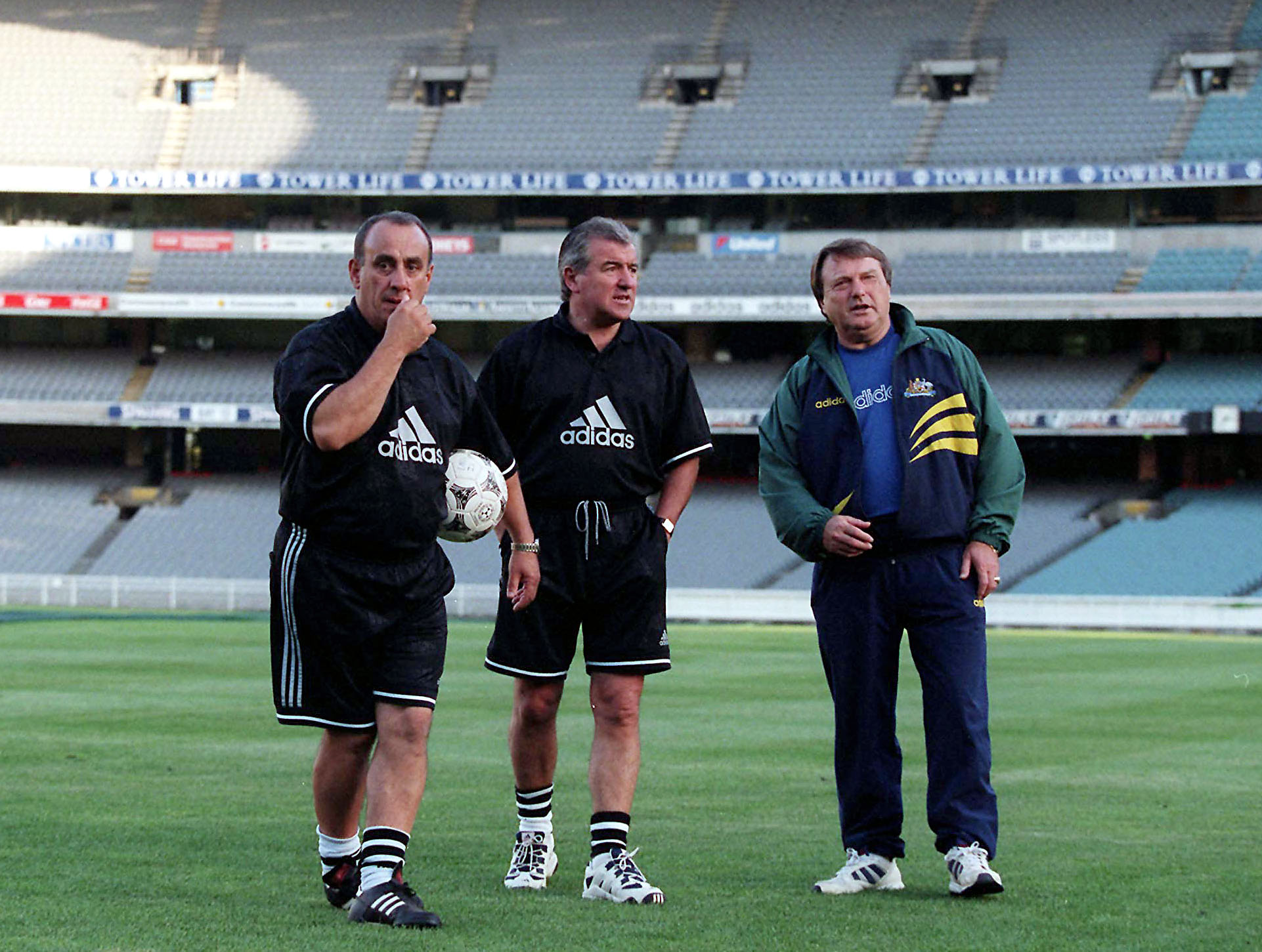 Three soccer coaches on a field inside an empty stadium.