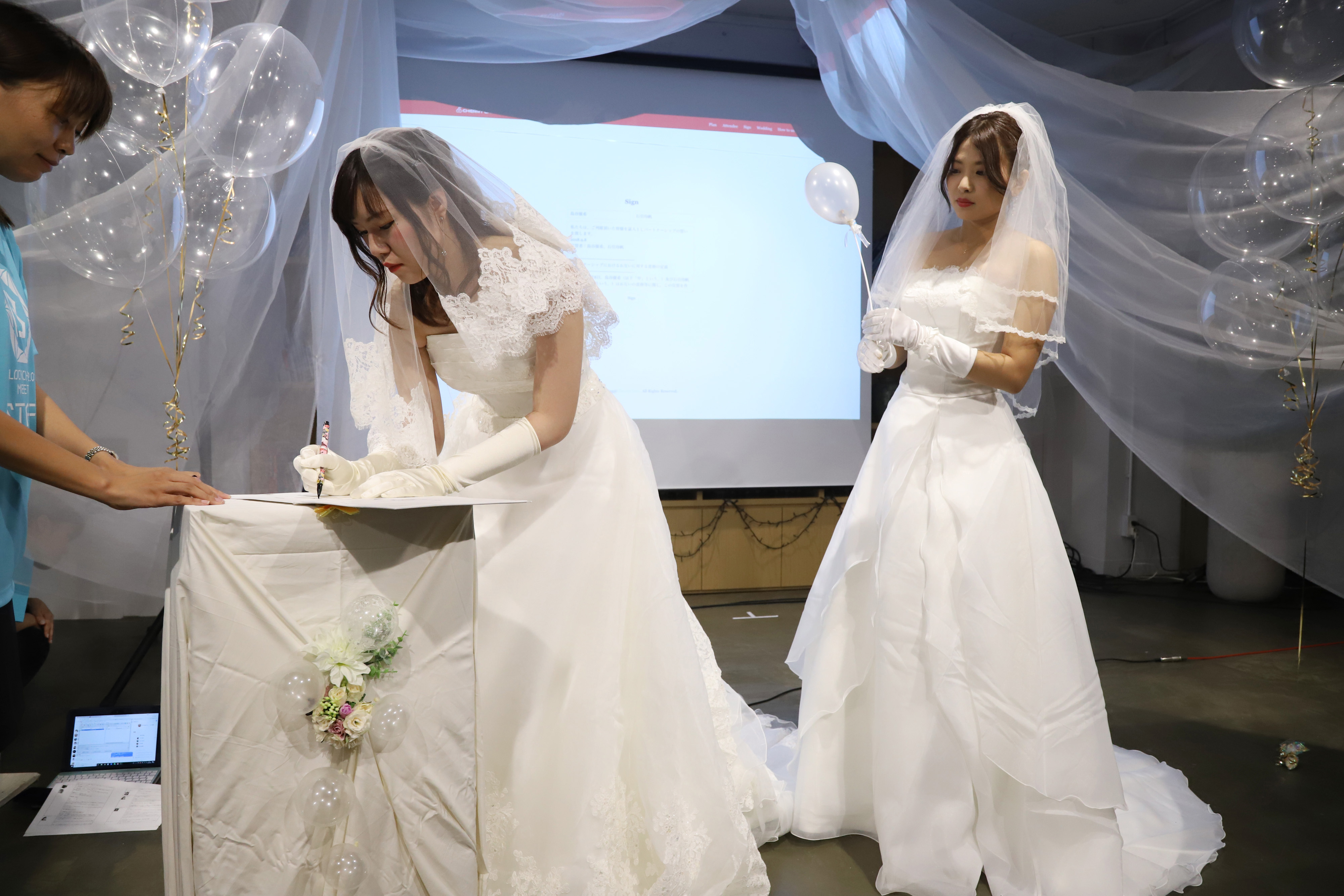 Two women in white bridal dresses in front of a projector screen, surrounded by clear balloons. One of the women is signing a wedding contract while the other watches with a balloon in hand.