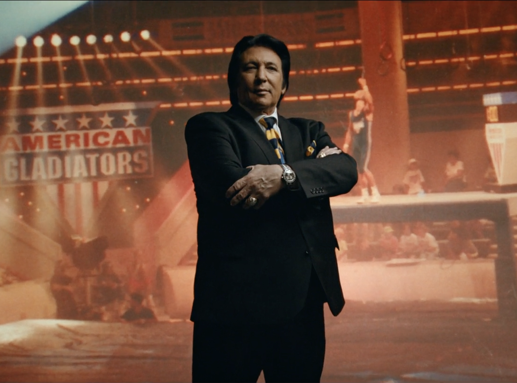 A man with black hair and wearing a suit stand in front of 'American Gladiators' wallpaper.