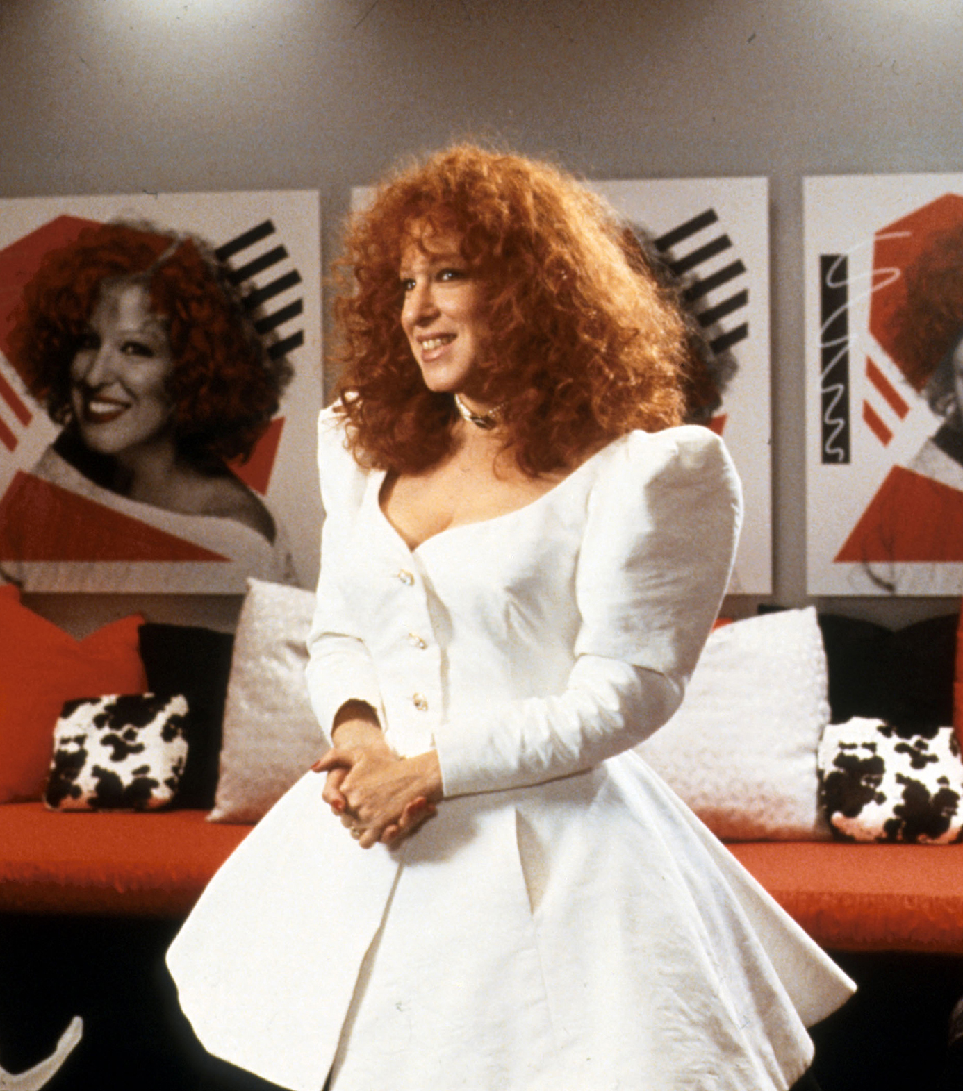 Bette Midler stands in a white dress with her hands held together. Portraits of her face line the walls behind her.