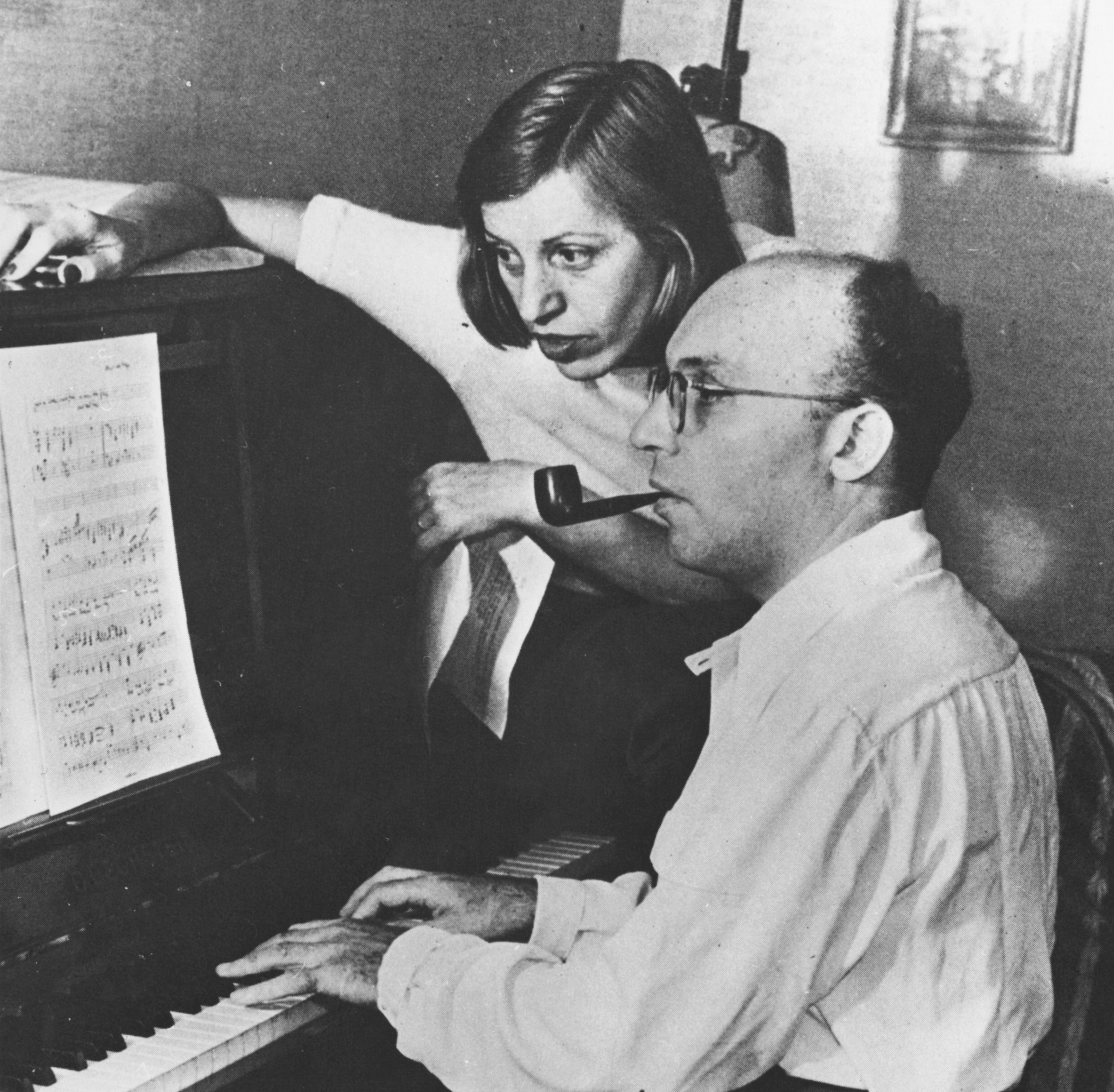 A balding man with glasses sits at a piano with a pipe in his mouth. A woman sits on the side of the piano, leaning in to read sheet music on the stand.