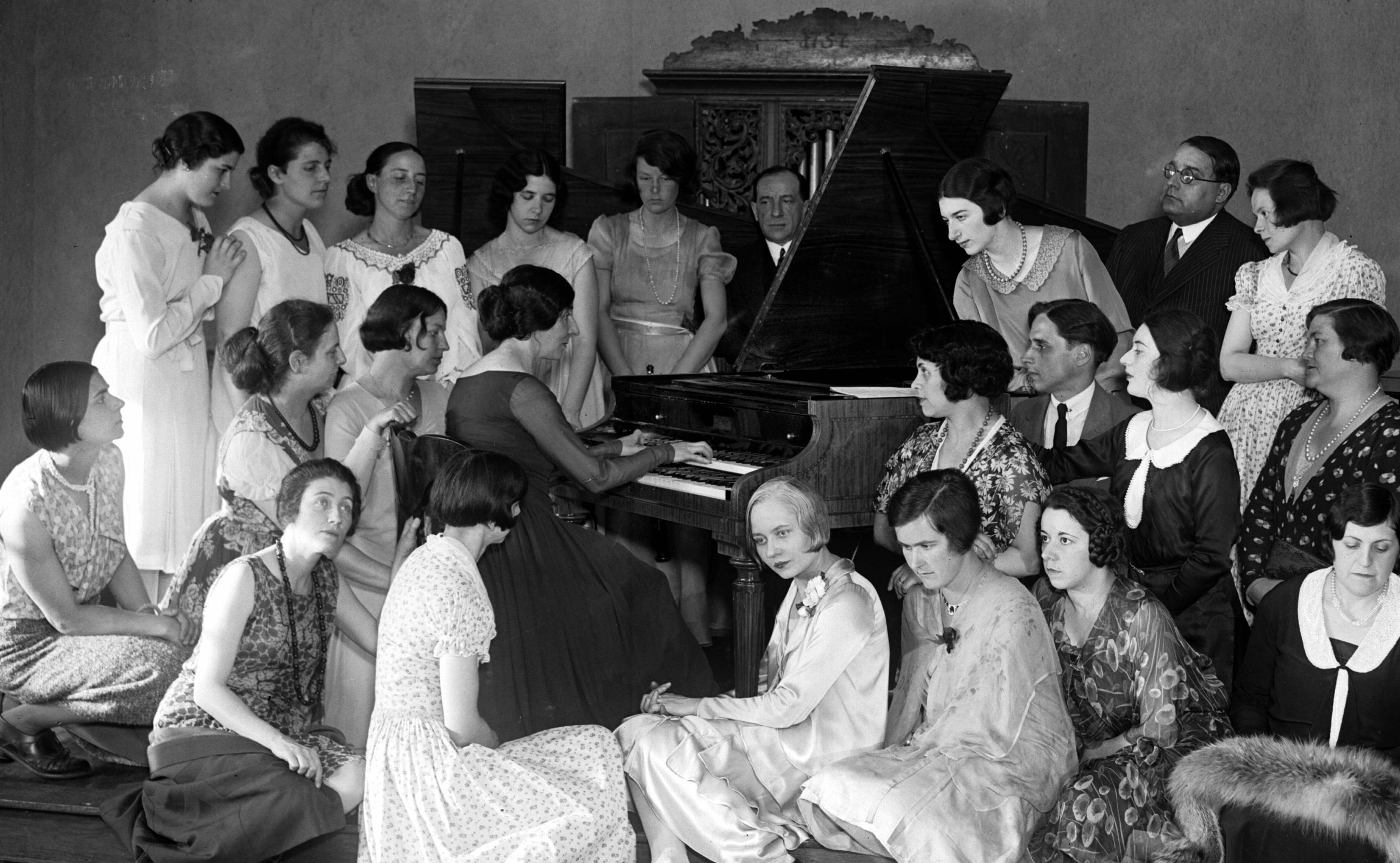 Black & white photograph of a woman playing a piano as she is surrounded by listening admirers.