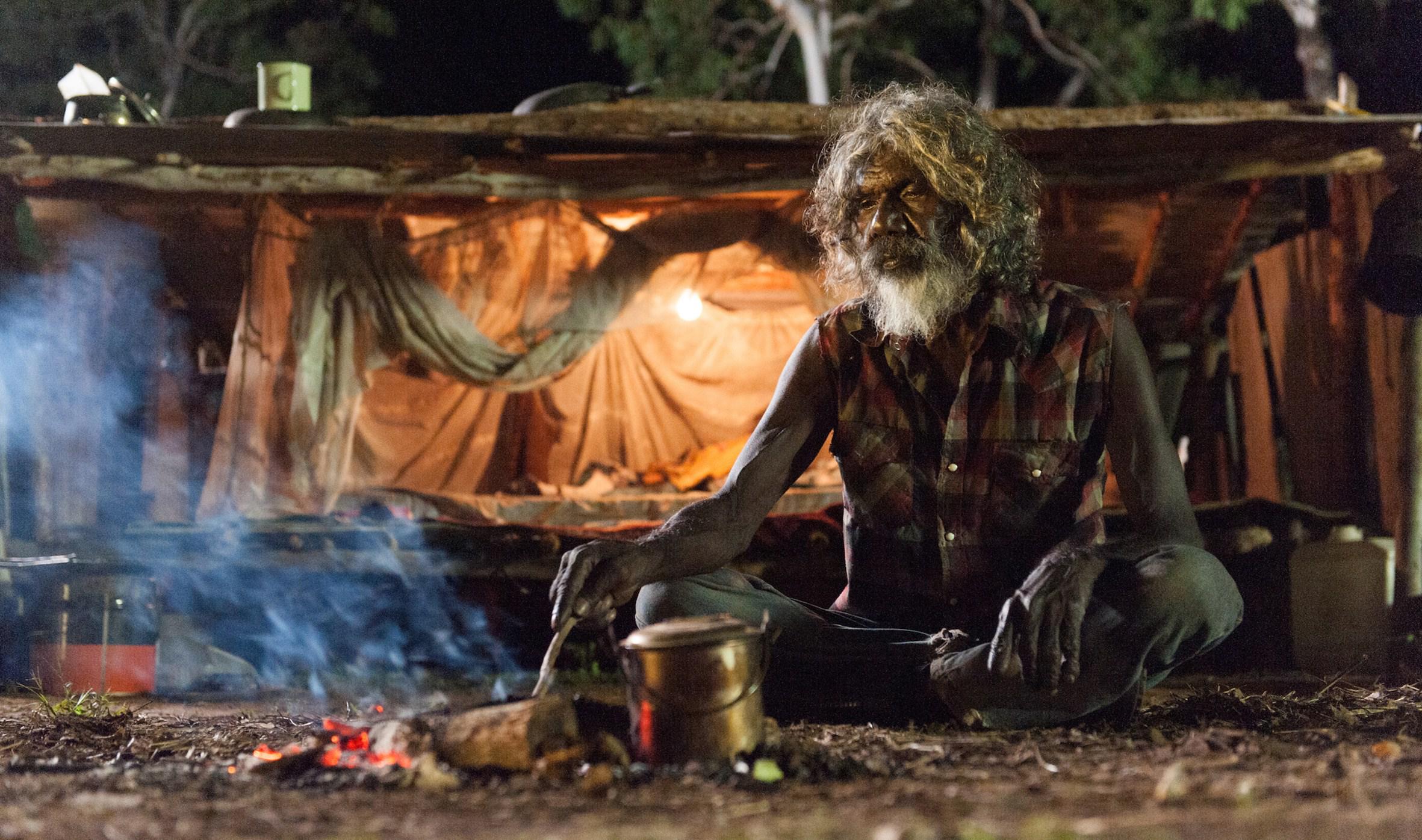 An elderly man with long hair and a grey beard sits by a campfire in the night, a lit up tent behind him