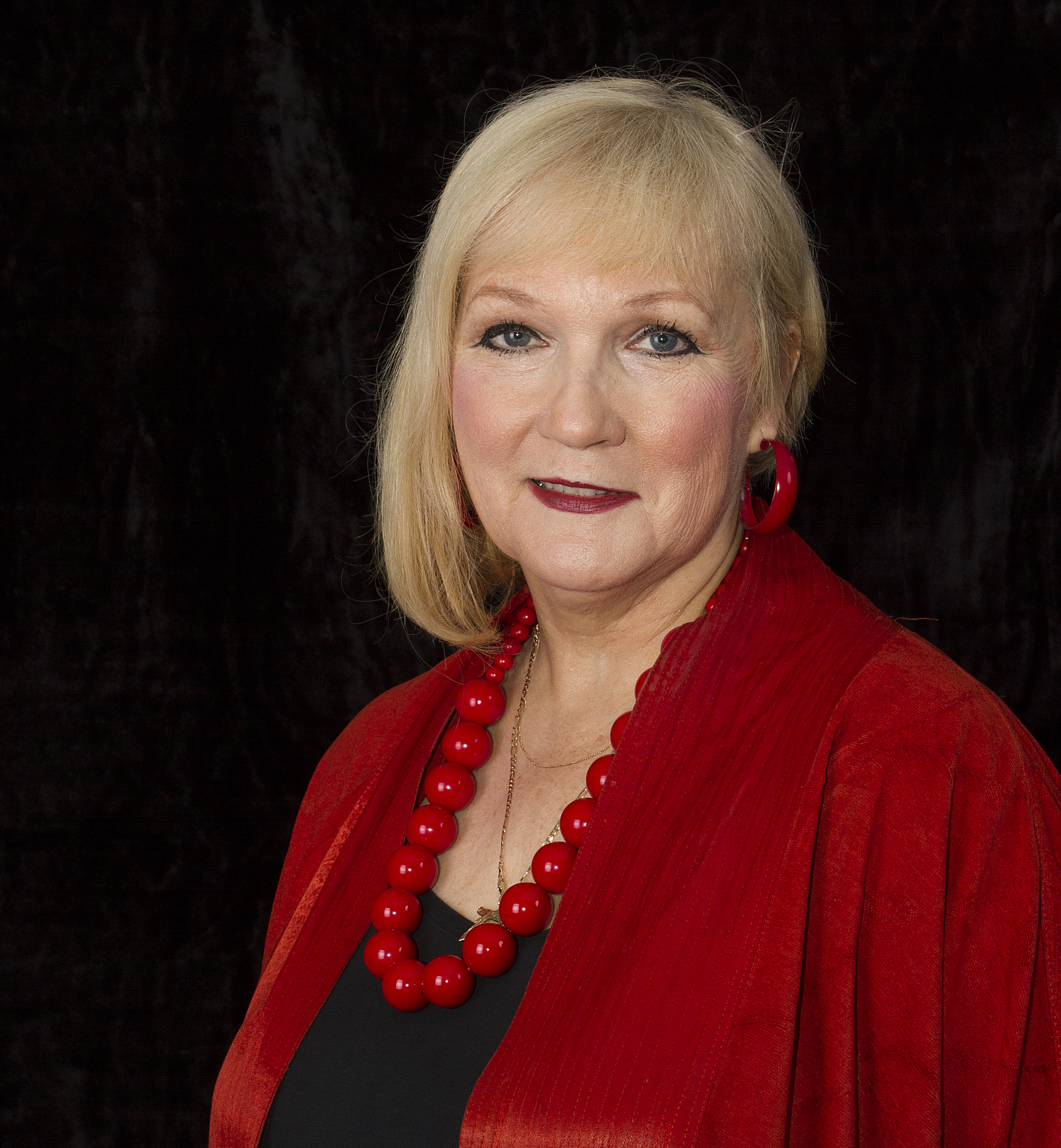 A portrait photograph of a woman with blonde hair, wearing a red dress, necklace and earrings.