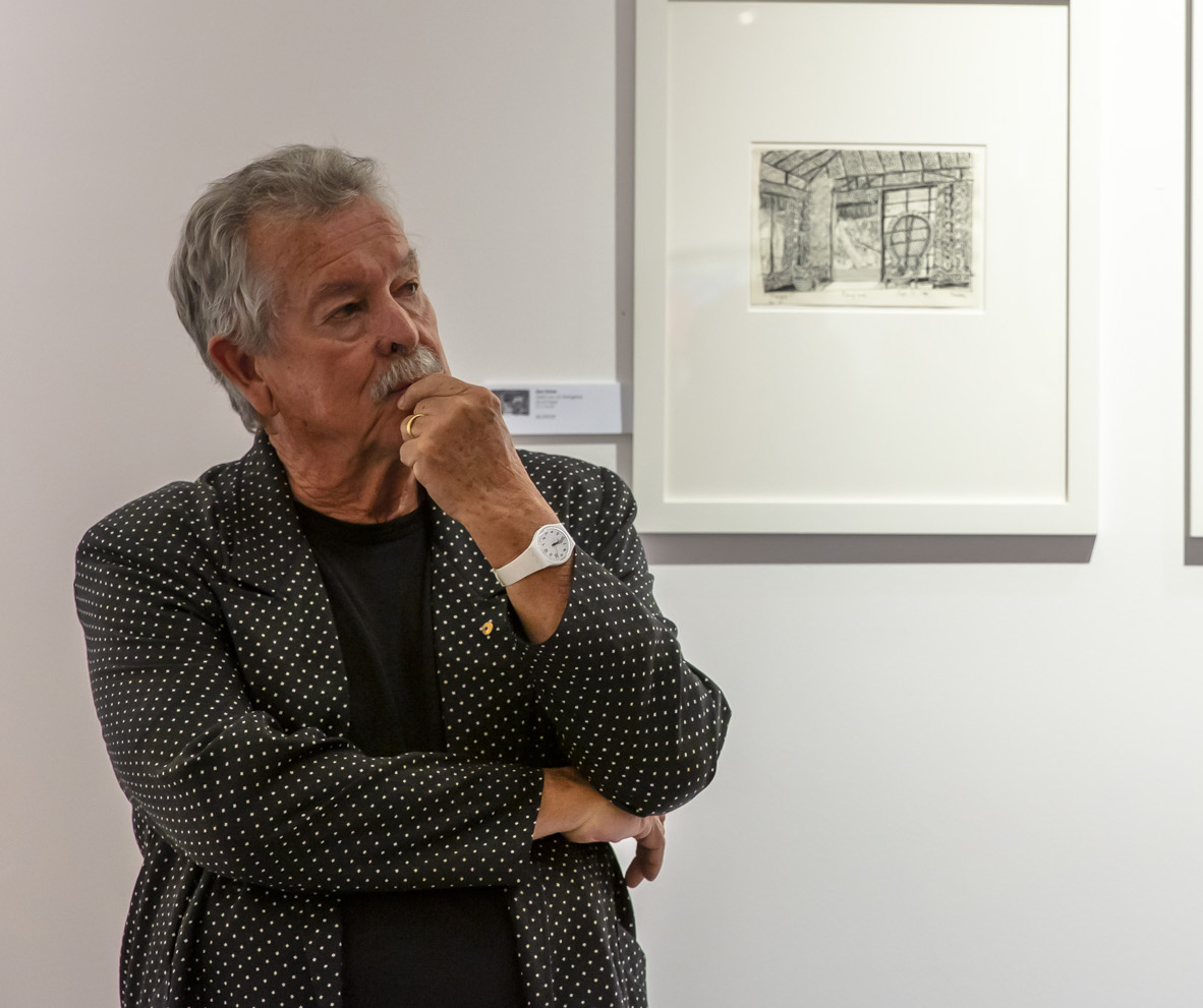 A man with a grey moustache and wearing a dotted blazer stands at an art exhibition with his ams crossed.