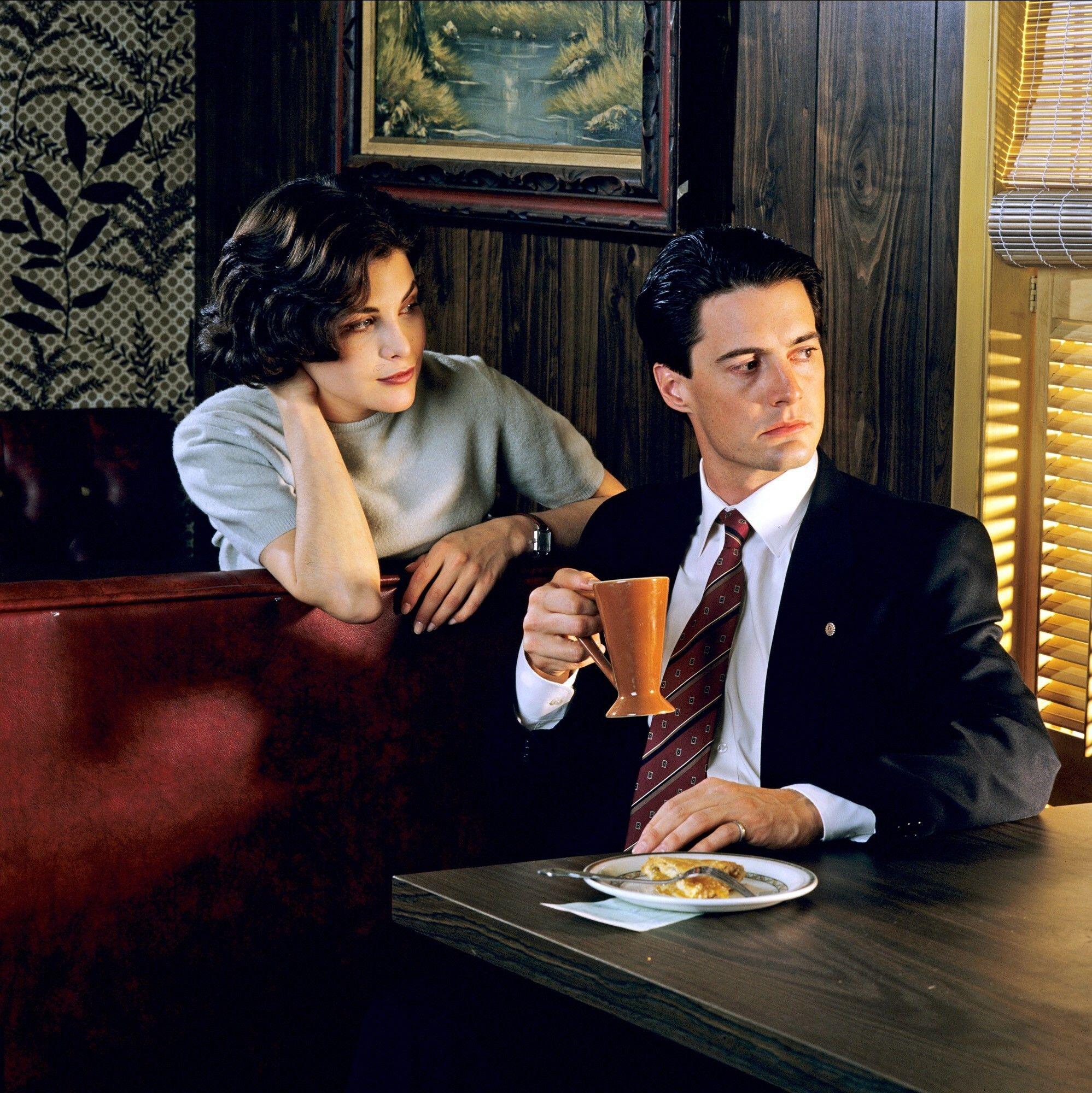 FBI agent Dale Cooper sits at a diner booth and gazes out the window, toward a bright light. Behind Dale, the young woman Audrey Horne is leaning over her own booth as she watches his face.