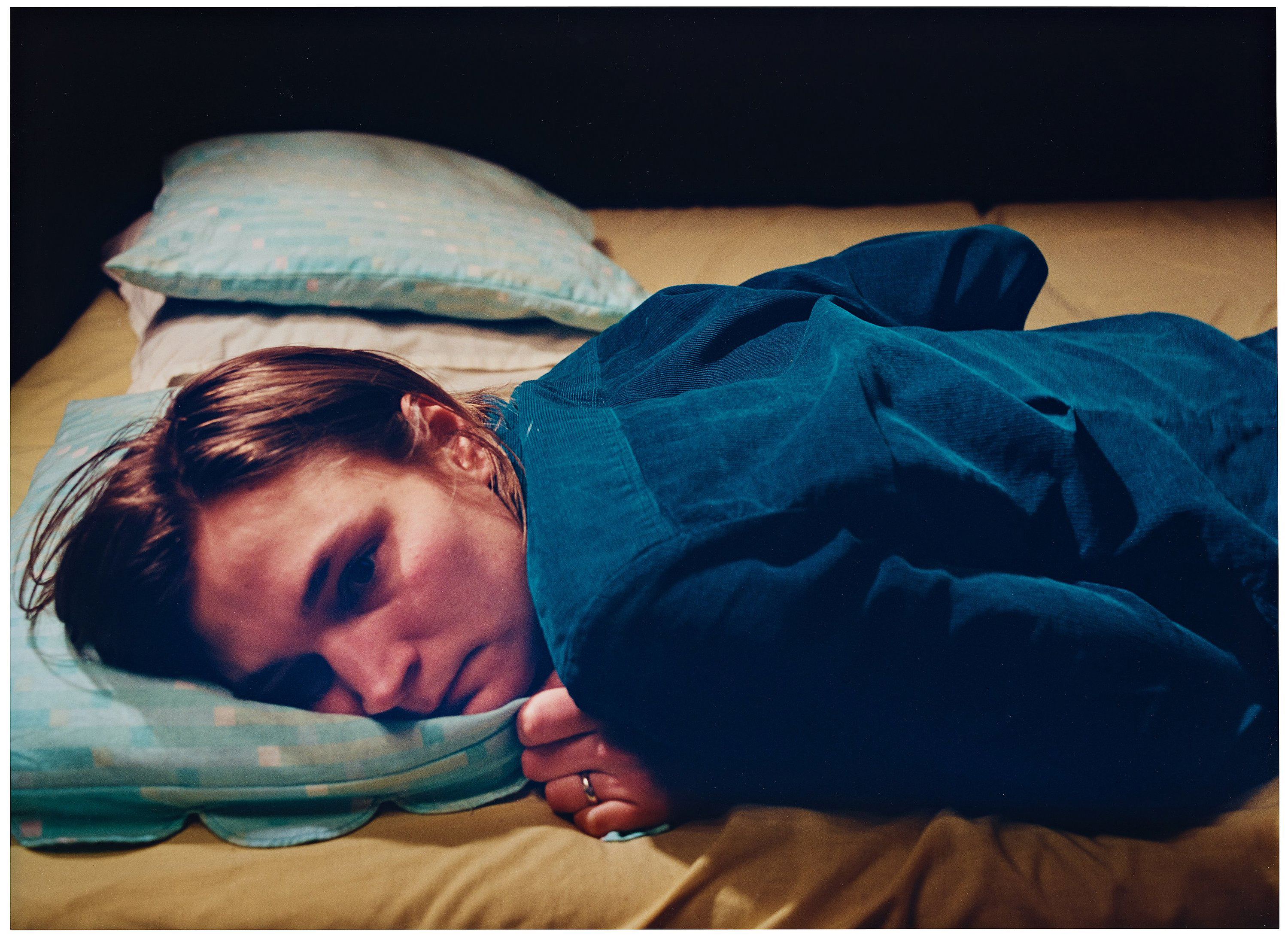 A moodily lit photograph of a woman with short hair lying belly-down on a bed, with her arms drawn up to her torso. Her face is empty and sad.